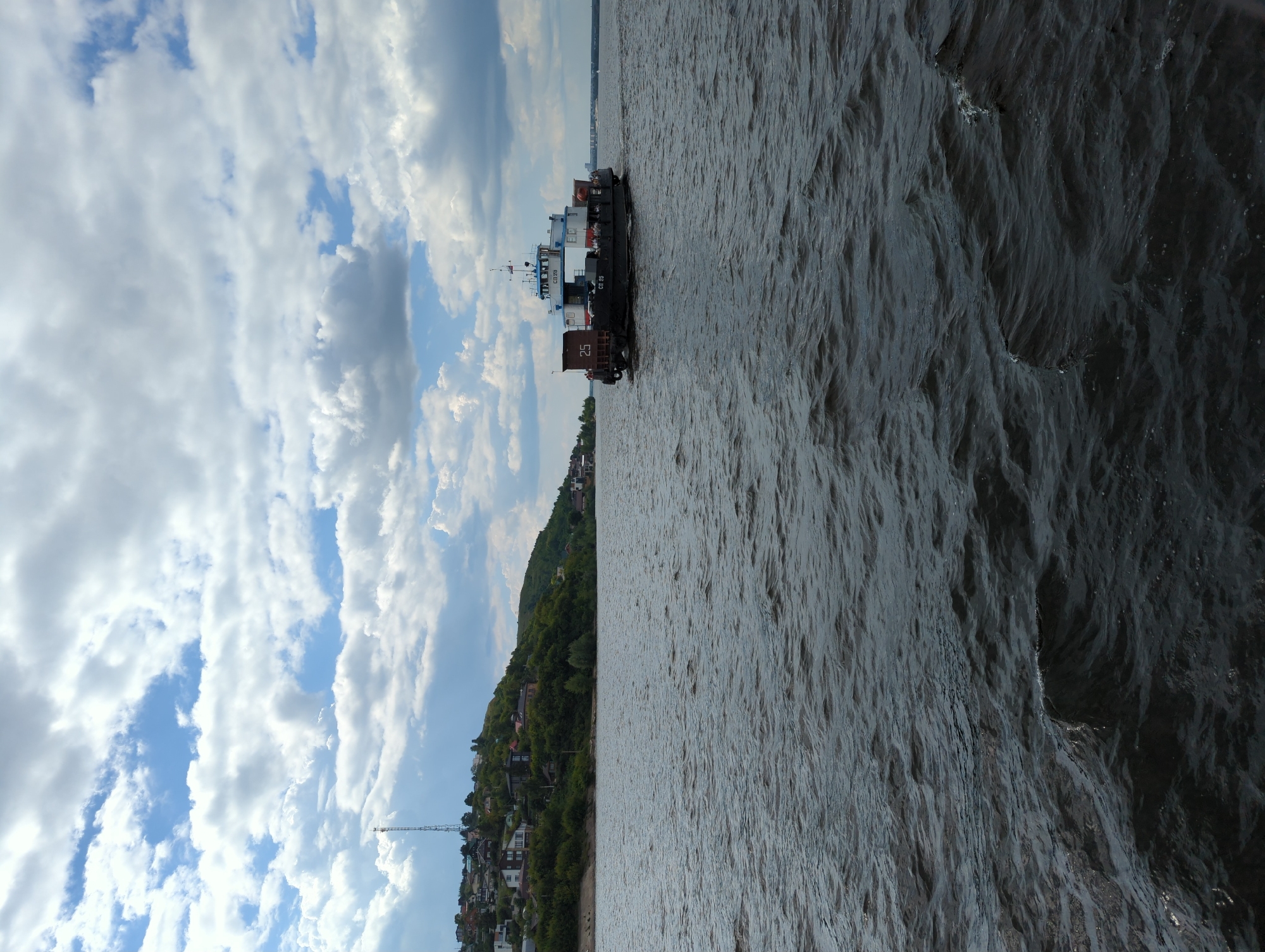 A ferry navigates a wide river under a cloudy sky. The ferry, painted dark gray with white upper structures and blue accents, carries several passengers and what appears to be cargo containers on its deck. The number "25" is visible on a dark rectangular object at the stern of the ferry. On the left bank of the river, a hill slopes upward, dotted with houses and trees. A tall antenna stands on the crest of the hill. The water's surface is choppy with visible ripples. The far bank of the river is distant and indistinct.