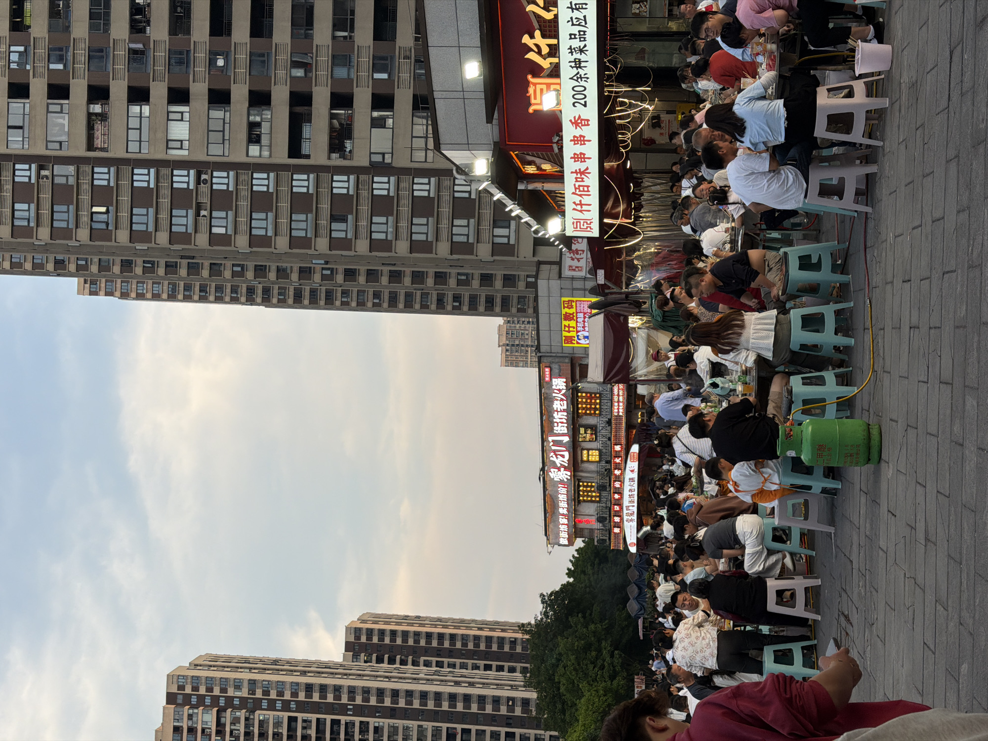 The image shows an outdoor dining scene in Chongqing High-tech Zone, China. Numerous people are seated on small, plastic stools at closely arranged tables, likely enjoying a meal at a local restaurant. Food stalls with illuminated signage are visible. A light green LPG (Liquefied Petroleum Gas) cylinder is present near one of the tables. Tall residential buildings rise in the background.