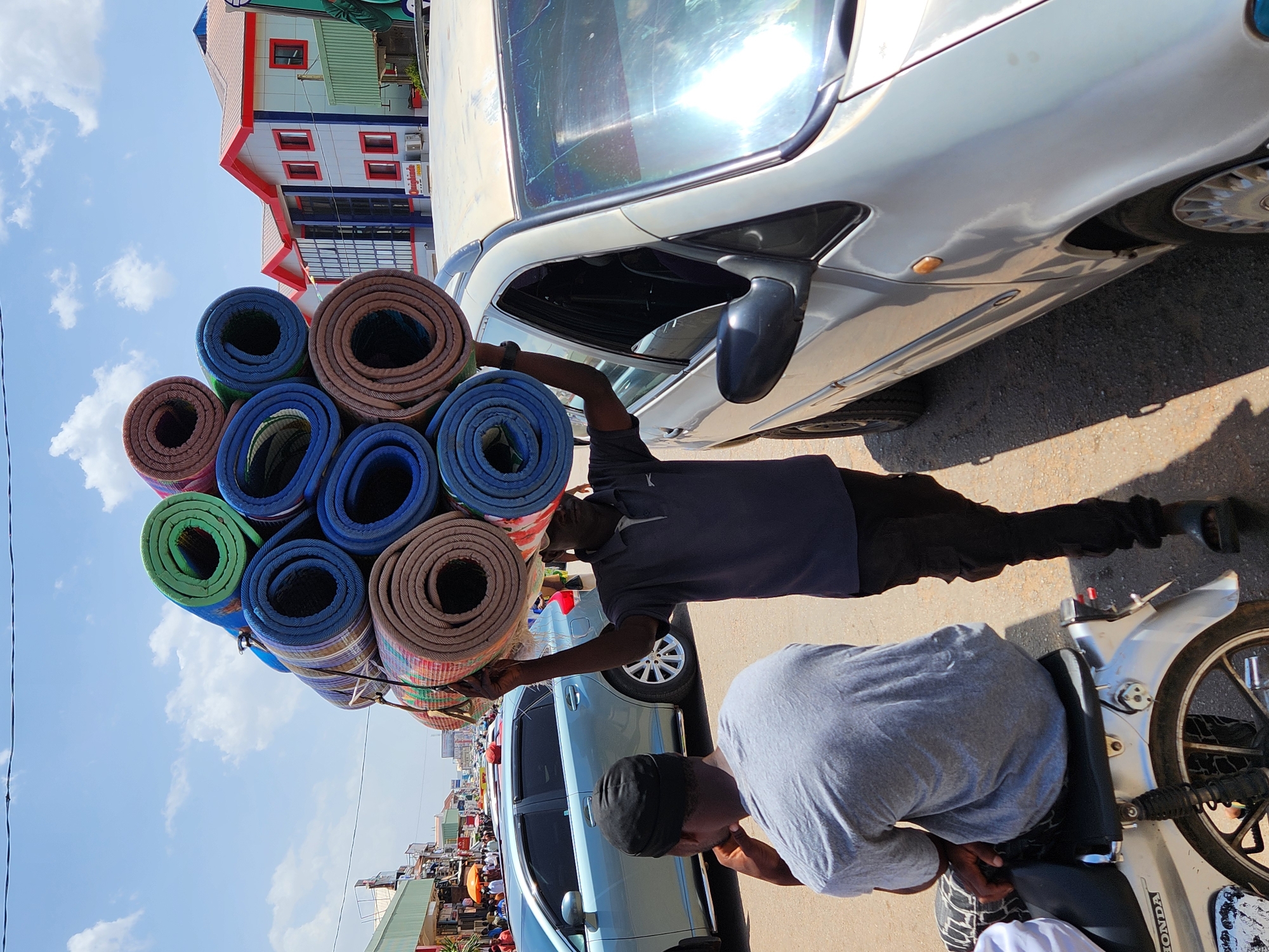 A daytime, eye-level shot shows two African men selling rolled mats to a car's occupants in an outdoor urban setting. The view is a street scene.
The seller holding the mats is wearing a dark gray short-sleeved T-shirt, dark pants, and black shoes. The second man is sitting on a white Honda motorcycle, wearing a gray T-shirt, black cap, and sneakers. He is bent over, looking down. The buyer is inside a silver sedan, only their arm visible to the seller.
Stacked high, the rolled mats are various colors, including green, blue, and brown. A building painted in red and white is visible behind the men on the left. A light blue sky is visible in the upper left of the frame. Several cars and pedestrians are in the background. The location appears to be a busy street in a city.