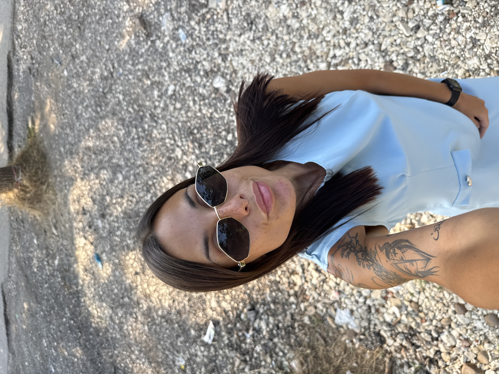 A woman with long dark hair and sunglasses is taking a selfie outdoors. She is wearing a light blue dress and has a tattoo on her left arm. The ground around her is covered in gravel and small stones, with some debris scattered about. The lighting suggests it is daytime. There are no specific landmarks visible that would allow for precise identification of a location within Pleven, Bulgaria.