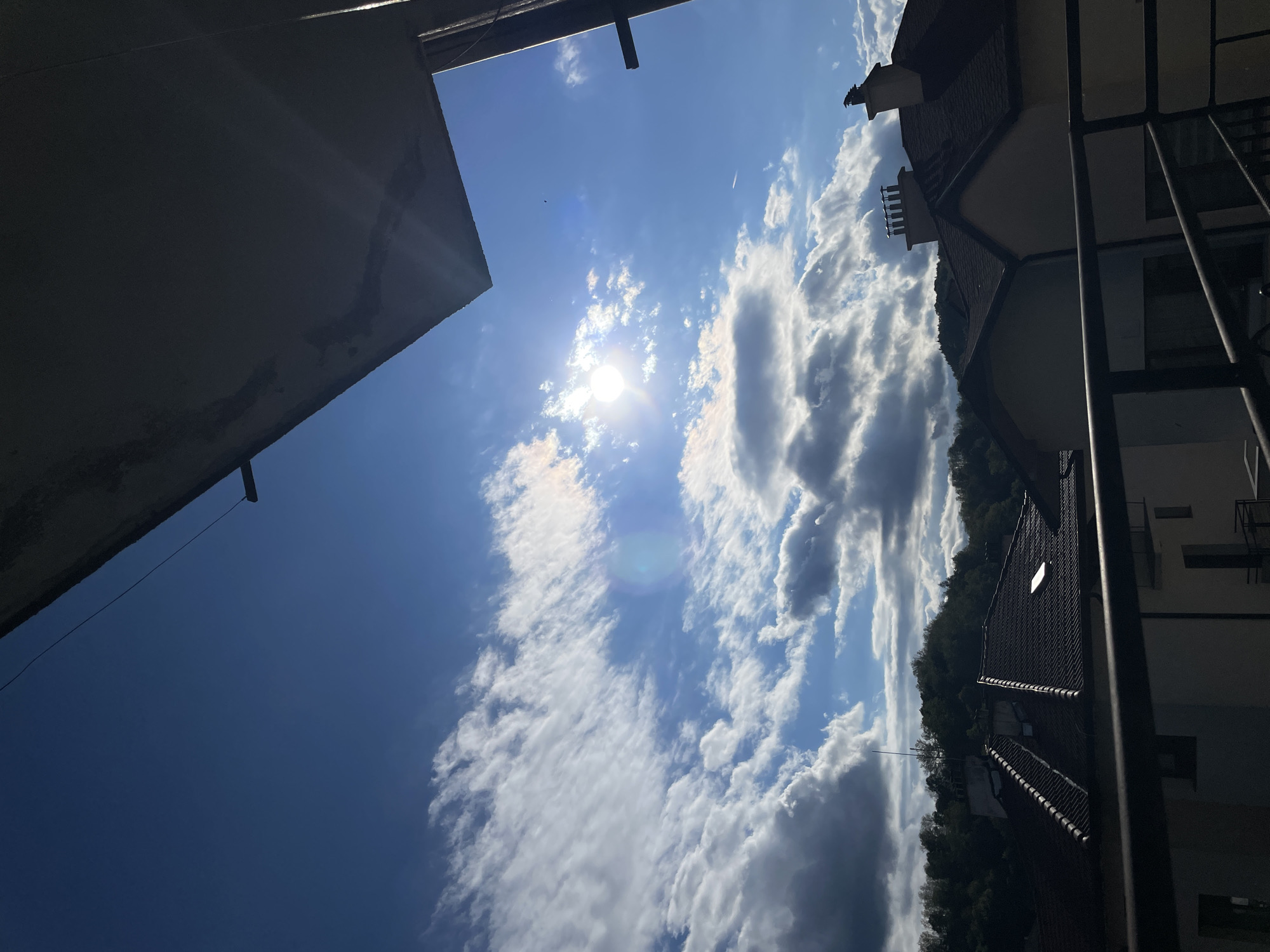 An upward-facing view captures a partly cloudy, bright sky. In the upper left, a building element in shadow partially obstructs the view. To the right, the roofline of a building is visible, complete with a chimney and other architectural details. Trees are visible in the background along the horizon. The location is likely Zlatograd, Bulgaria, given the prompt.