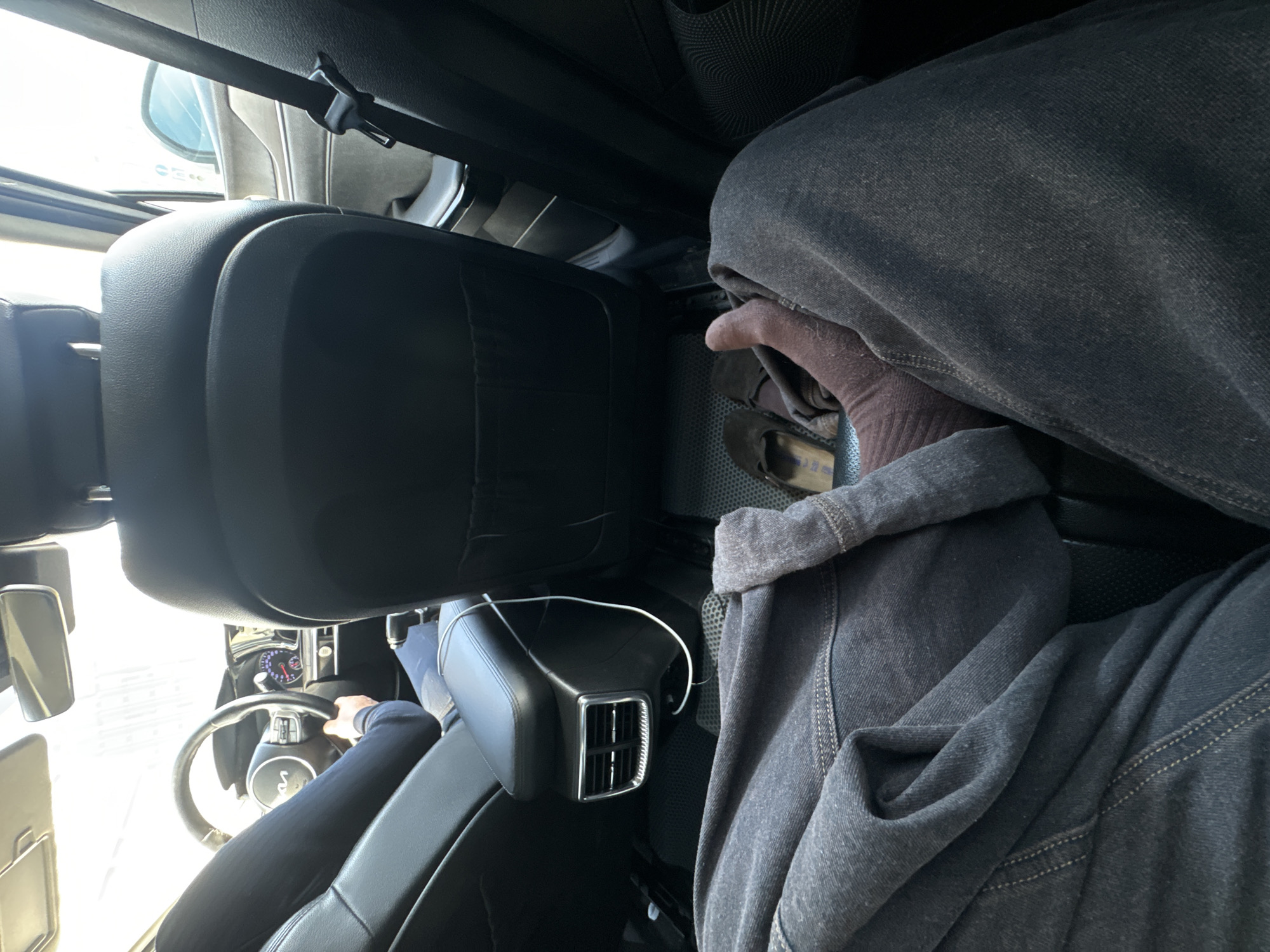 The image is taken from inside a car, presumably in the back seat. A person wearing a gray hooded jacket is present in the shot. A portion of a steering wheel and dashboard can be seen through the gap between seats in the foreground. It is impossible to verify the location of Moscow, Russia, based on the context of this image.