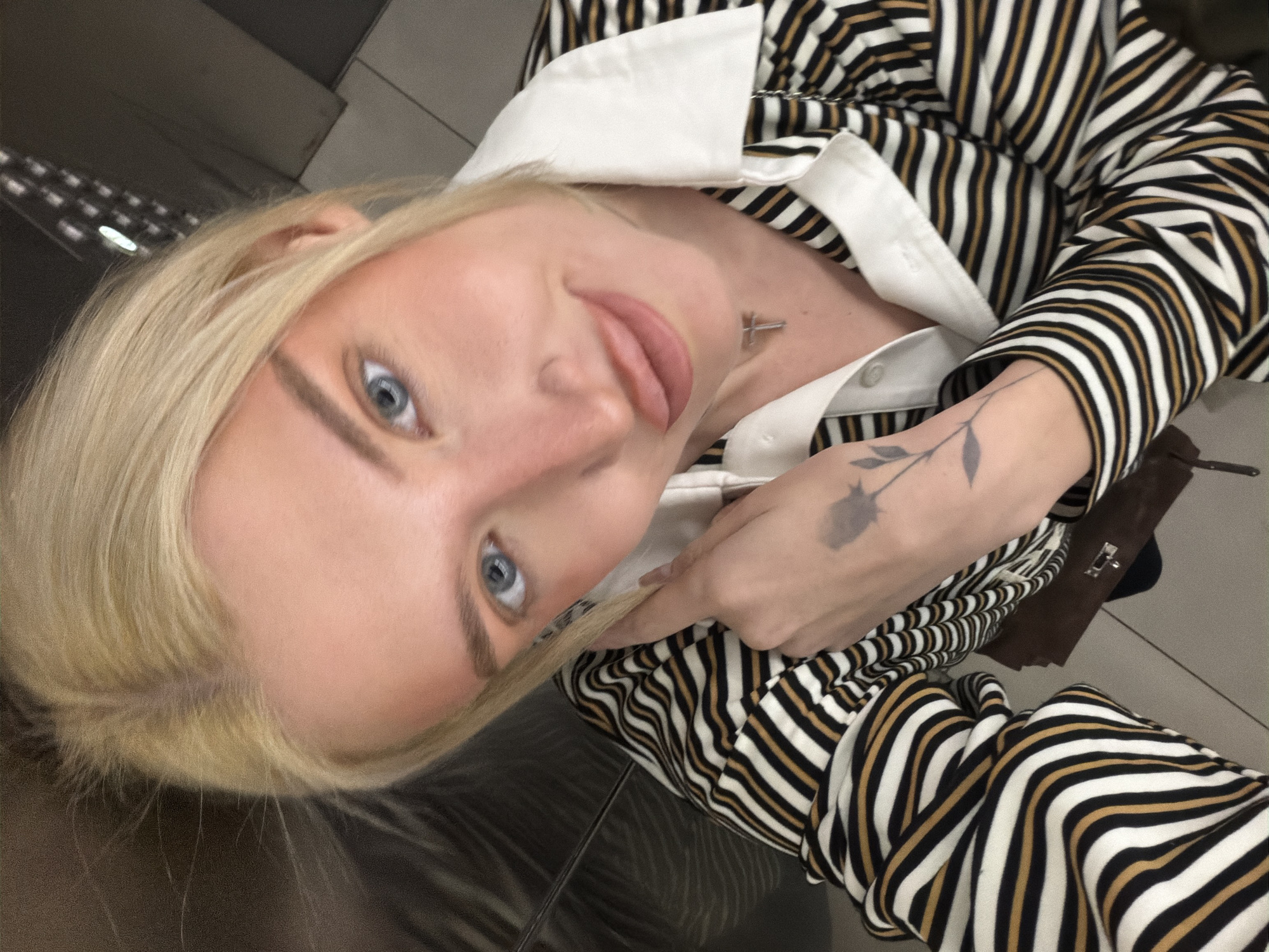 A woman with blonde hair and blue eyes is taking a selfie. She is wearing a striped shirt with a white collar and has a tattoo of a flower on her forearm. She appears to be indoors, possibly in a building with a tiled floor. The location in Belgrade, Serbia is not discernible from the image.