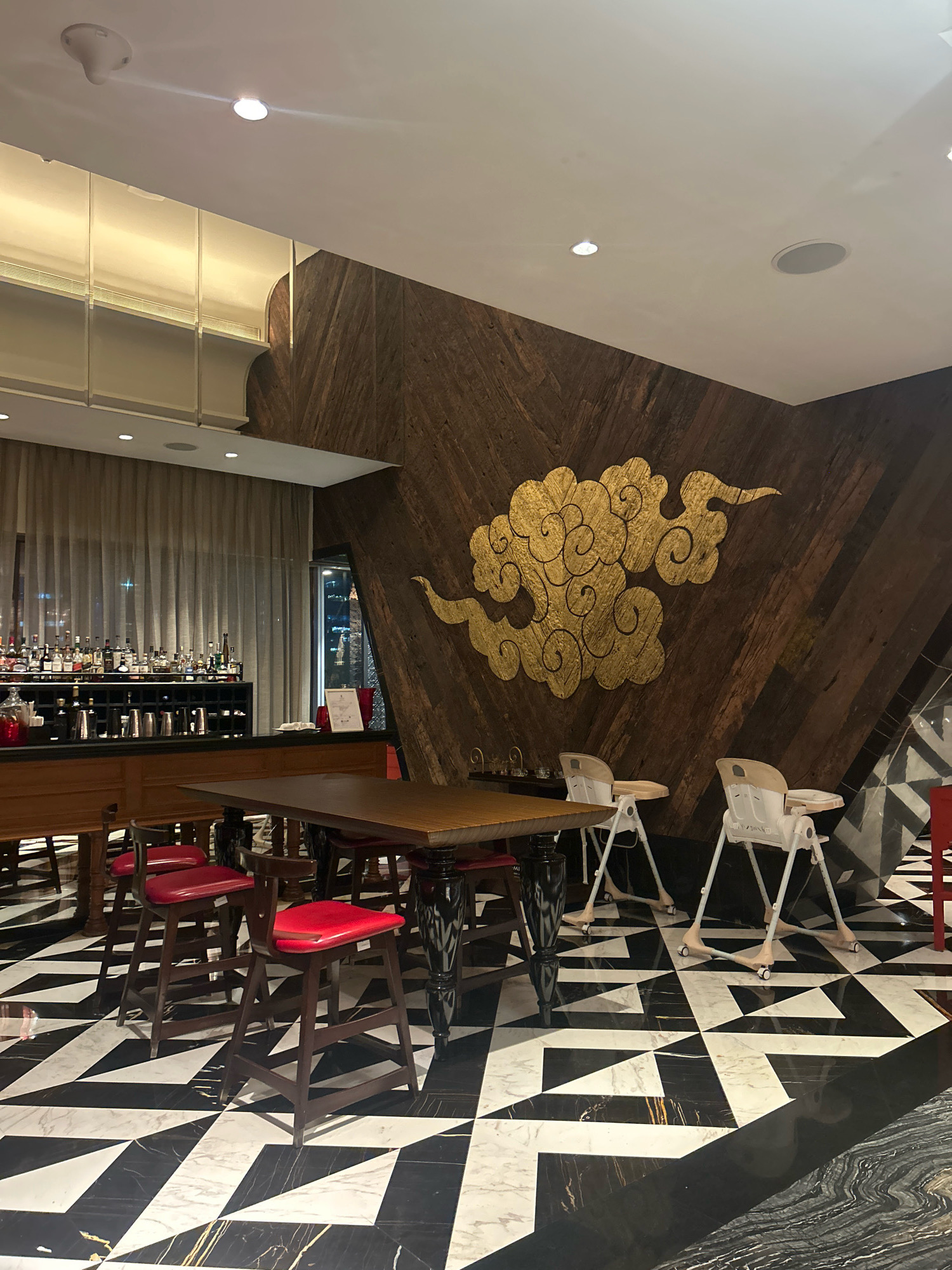The image depicts an interior scene of a restaurant or bar. The floor has a striking black and white geometric pattern, transitioning to a marble pattern on the right. A wooden table with a dark, ornate base is centrally positioned, surrounded by dark wooden bar stools with red seats. Two white high chairs are placed behind the table. The wall behind the table is decorated with a large, ornate gold cloud design on a dark wooden paneling. To the left, a bar area is visible with a counter, numerous bottles of liquor on shelves, and a draped beige curtain. The lighting is dim and ambient, with some spotlights illuminating the ceiling. The overall aesthetic suggests a modern and stylish establishment. The location in Hyderabad, India, is not discernible from the visual elements present in the image.