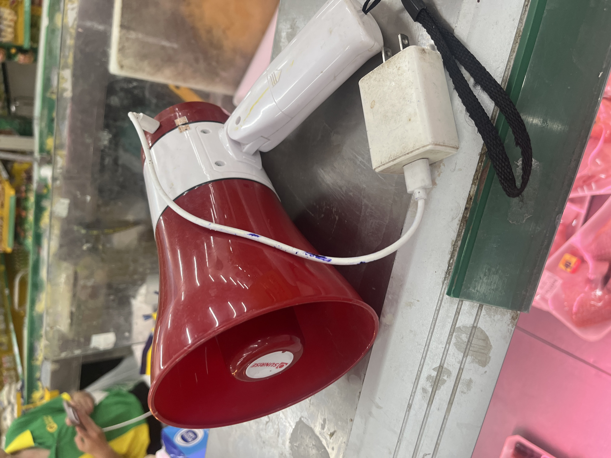 A red and white megaphone is resting on a metal surface. A white charging cable is plugged into the megaphone and connected to a white electrical adapter. The adapter is also plugged into an outlet. A black wrist strap is attached to the adapter. In the background, a person wearing a green and yellow shirt is holding a phone. The scene appears to be in a store, possibly a supermarket, given the presence of shelves and products in the background. The lighting is bright, suggesting an indoor environment.