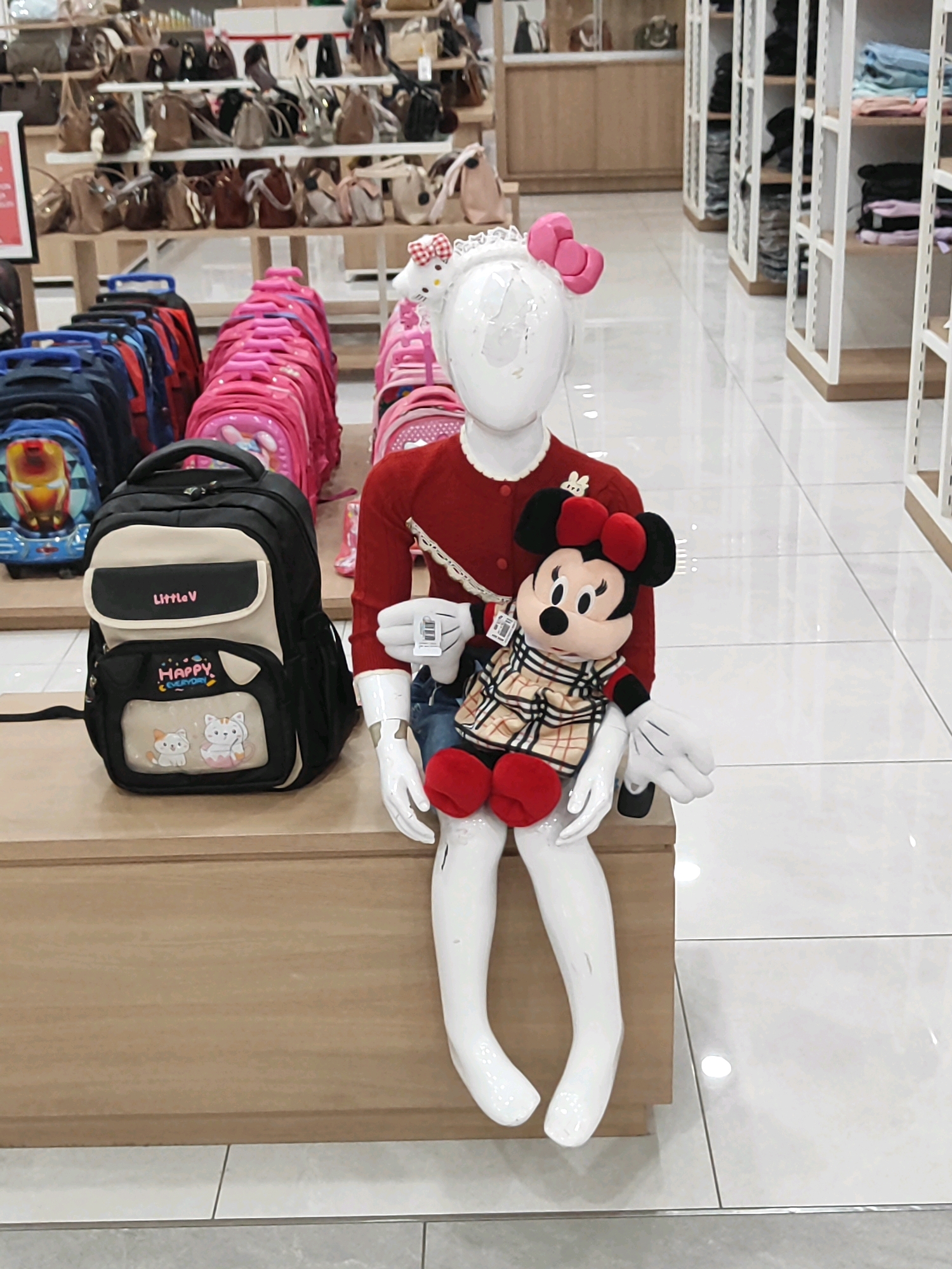 The image shows a retail environment. In the foreground, a white mannequin is seated on a wooden display. The mannequin is dressed in a red sweater and blue jeans, and is holding a Minnie Mouse stuffed toy. To the left of the mannequin is a black backpack with a beige and black front pocket that reads "Little V" and has an illustration of two cats. Behind the mannequin, several colorful backpacks are displayed on a shelf. In the background, shelves are filled with various handbags and other merchandise. The floor is tiled and appears to be a polished white. The scene is well-lit, suggesting an indoor retail space. This scene is not specifically located in Sukowiryo, Indonesia, nor does it contain any elements that would identify it as being in that location.