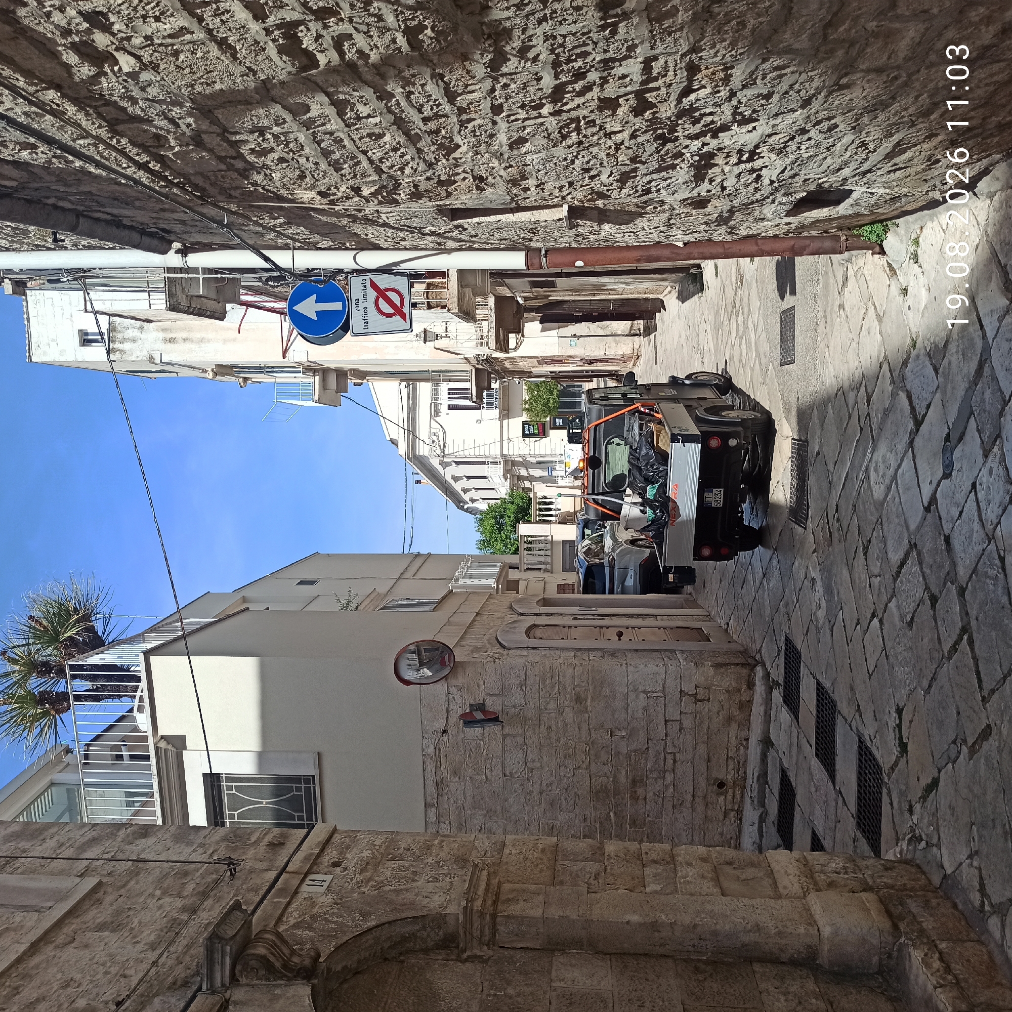 This image captures a narrow, sun-drenched street in Ruvo di Puglia, Italy, with buildings on either side constructed from rough-hewn, light-colored stone. The street is paved with irregular flagstones, and the buildings feature balconies and windows. On the left, a stone archway is visible. A small, dark utility vehicle with a cargo bed filled with black bags is parked in the middle of the street. The vehicle has a license plate that reads "WF 58234". Traffic signs are present: a blue circular sign with a white arrow pointing up is mounted on a pole, accompanied by a rectangular sign below it with Italian text and a red circle with a diagonal line through it. A convex mirror is attached to the wall on the left. The sky is clear and blue. In the background, more buildings and some greenery are visible. A date and time stamp, "19.08.2026 11:03", is overlaid on the bottom right of the image.