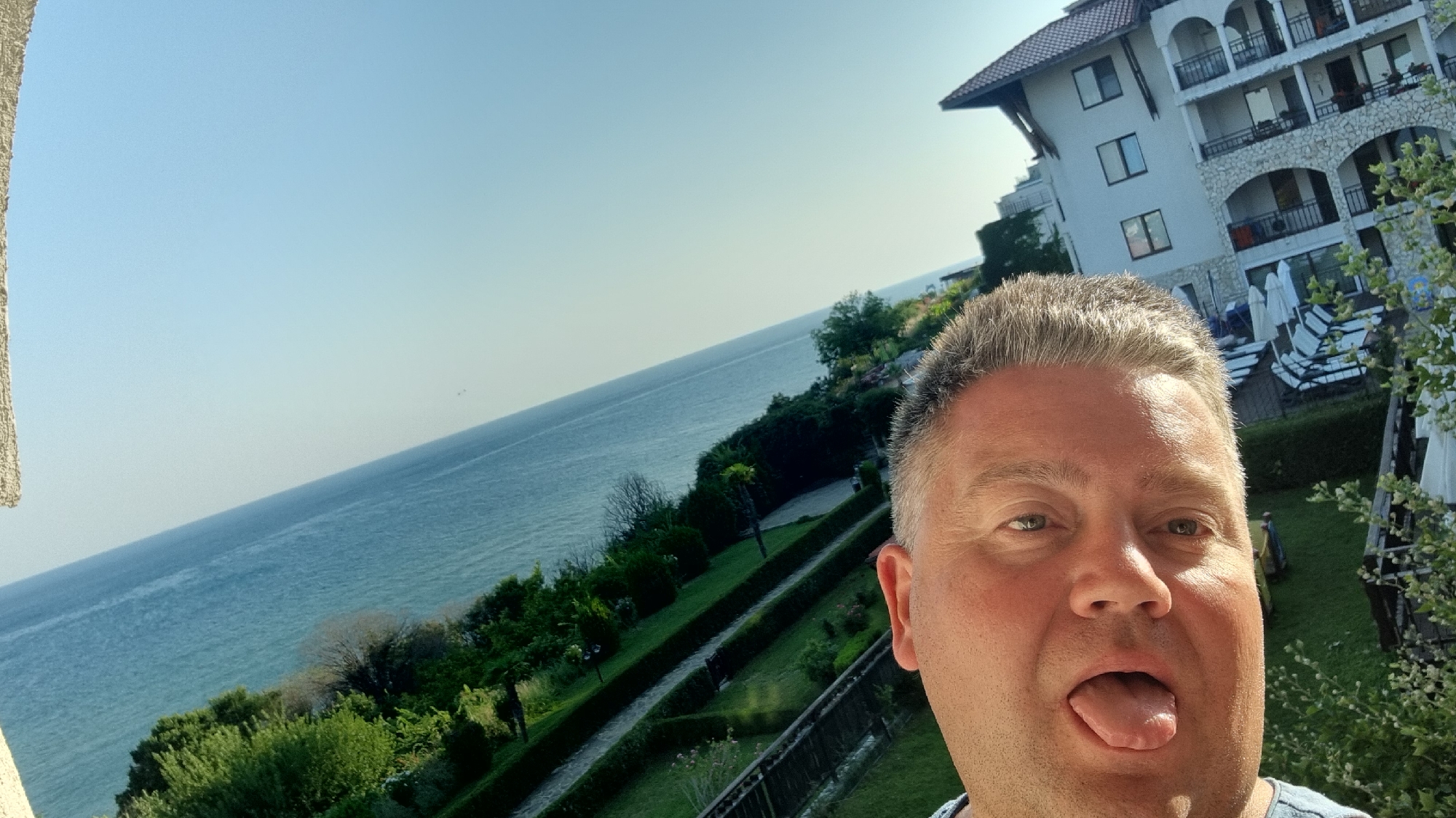 This is a selfie taken by a man in Sveti Vlas, Bulgaria. He is in the foreground, sticking his tongue out at the camera. Behind him, there is a white building with balconies and arched windows, and a view of the Black Sea and lush greenery. The man appears to be on a balcony or terrace, looking out at the scenic vista. The building is characteristic of resort architecture in the area, suggesting a vacation setting. The sky is clear and blue, indicating good weather.