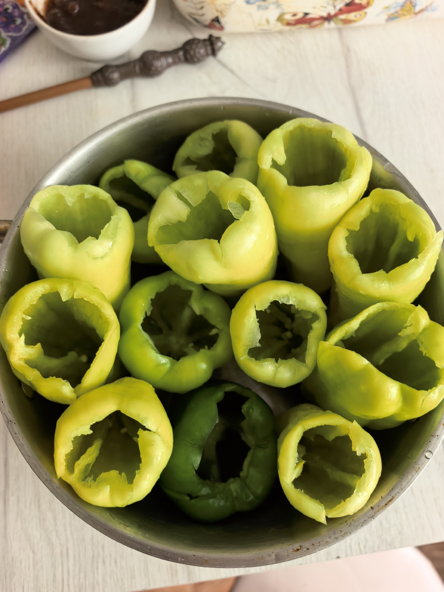 A top-down, close-up shot captures a silver pot filled with hollowed-out green bell peppers. The peppers are of varying shades of green, with some appearing lighter yellow-green. Their tops have been cut off, revealing their hollow interiors, and they are arranged in a somewhat disorganized manner within the pot. The pot is placed on a light-colored, wooden surface, possibly a table. In the background, a small white bowl containing a dark substance is visible, alongside a decorative item that resembles a wand. The overall impression is of ingredients prepared for a cooking or culinary activity. There is no indication of Sterlitamak, Russia in the image.