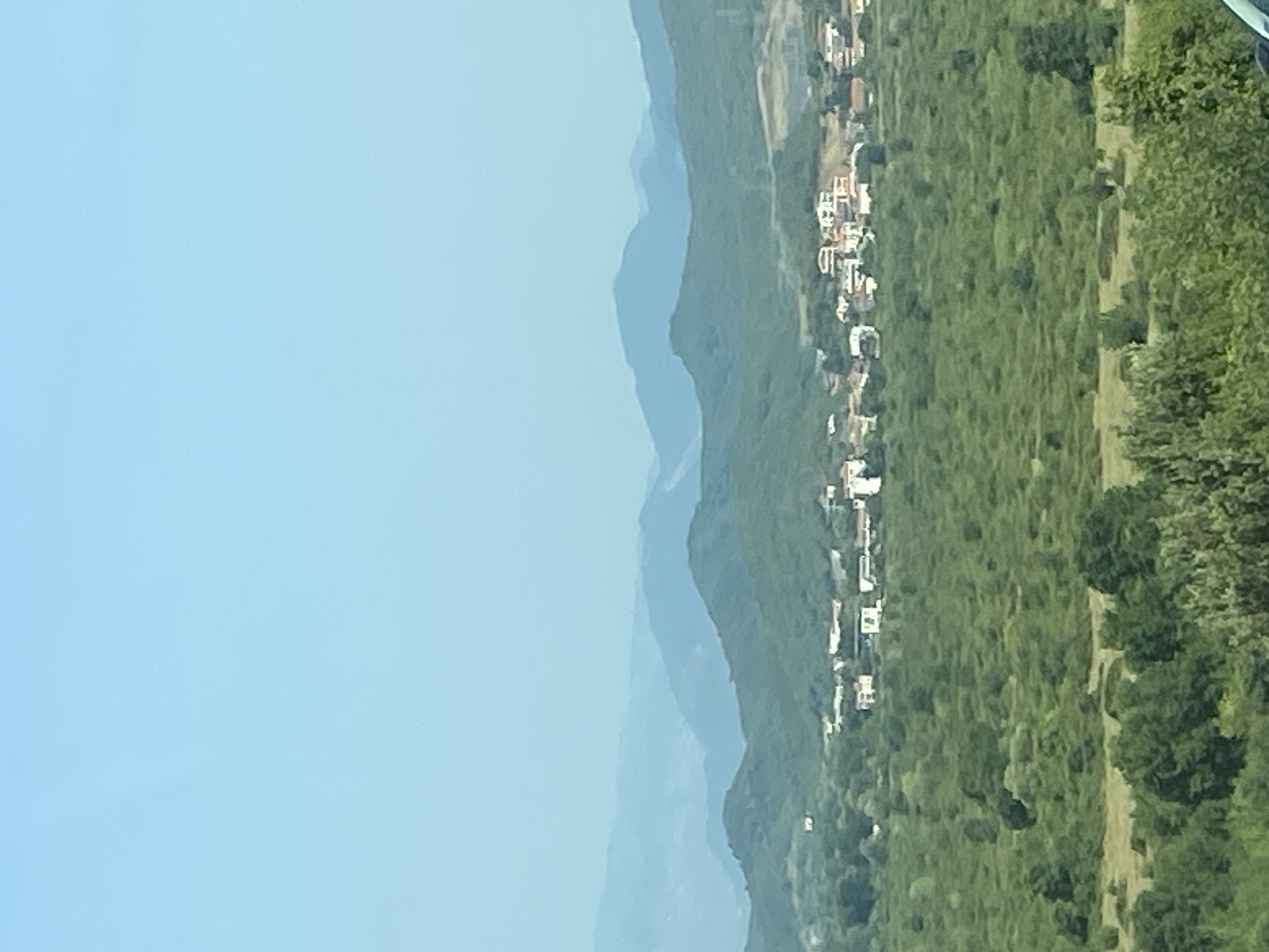 The image shows a panoramic view of a rural landscape with a town nestled amongst green hills and mountains under a clear blue sky. In the foreground, lush green foliage and trees are visible. The middle ground features a densely vegetated area, dotted with trees. Beyond this, a cluster of buildings, characteristic of a small town, is situated on a gentle slope. These structures appear to be houses and other buildings with red-tiled roofs and white walls. In the background, successive layers of green and blue mountains recede into the distance, suggesting a vast and undulating terrain. The overall scene conveys a sense of tranquility and natural beauty. The image is static, showing a typical day in a remote, scenic location. It is not possible to identify specific people or actions as the focus is on the landscape and the settlement. The location is identified as Prijevor, Montenegro.