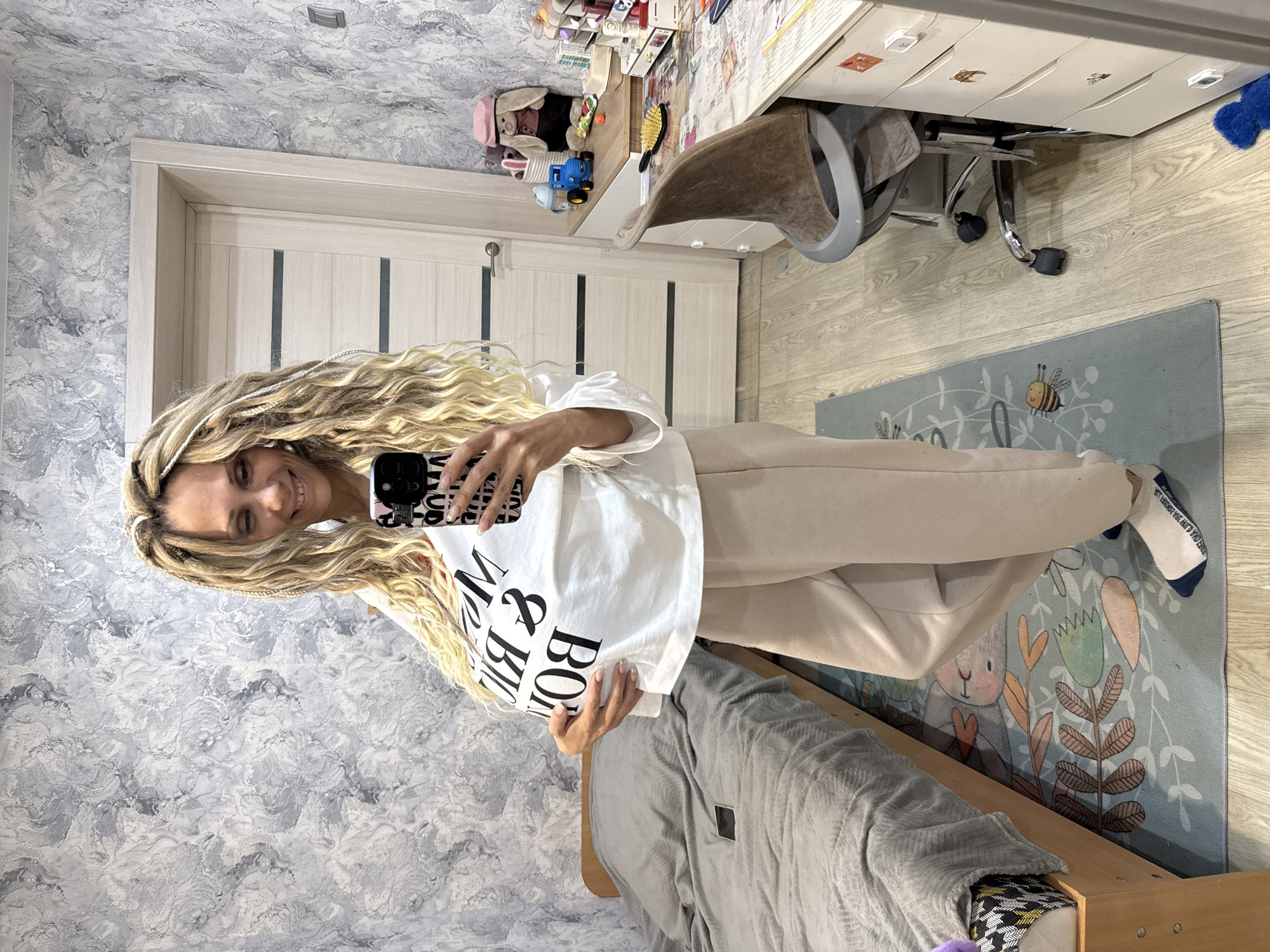 A woman stands in a room, angled to face a mirror and taking a selfie. She has long blonde dreadlocks and is wearing a white t-shirt with black lettering and tan pants. She is smiling and holding a black smartphone. Behind her is a bed with grey bedding, and a blue rug is visible on the floor. A door and textured grey and white wallpaper can be seen in the background. In the distance, a desk with drawers and an office chair are visible. There is no information available in the image to confirm the location as Rusanovo, Russia.