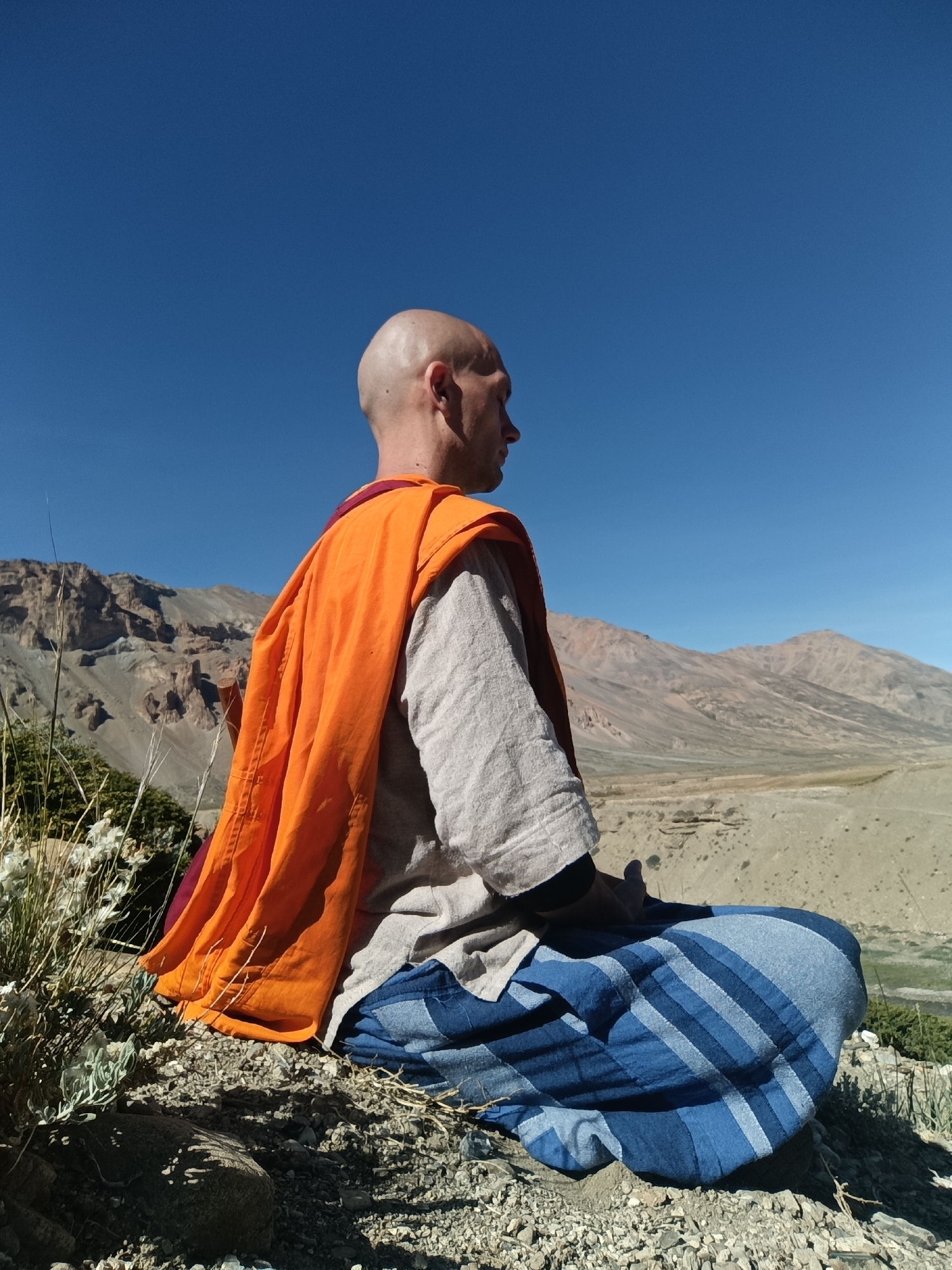 A bald man in Buddhist monk's attire, an orange shawl draped over his shoulders and a blue-striped cloth wrapped around his legs, sits in a meditative pose facing away from the camera. He is positioned on a rocky, uneven surface, with sparse, dry vegetation growing around him. In the background, a vast, arid landscape unfolds, characterized by rugged, barren mountains under a clear, deep blue sky. The location is Sarchu, India, a remote, high-altitude region known for its dramatic, desolate scenery.