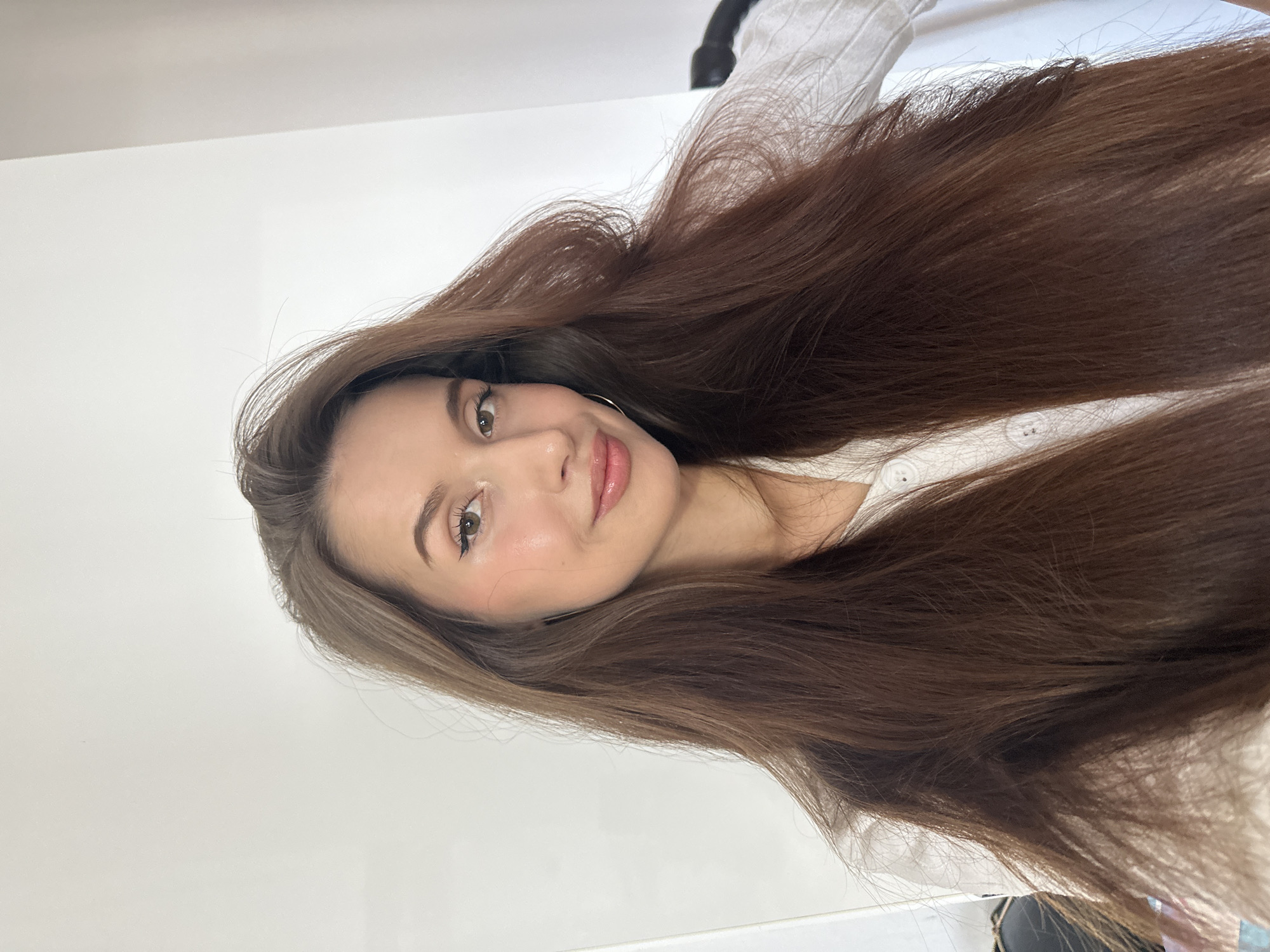 A woman with long, brown hair poses indoors. The background is plain white. She wears a white shirt. The location in Yekaterinburg, Russia is unknown.
