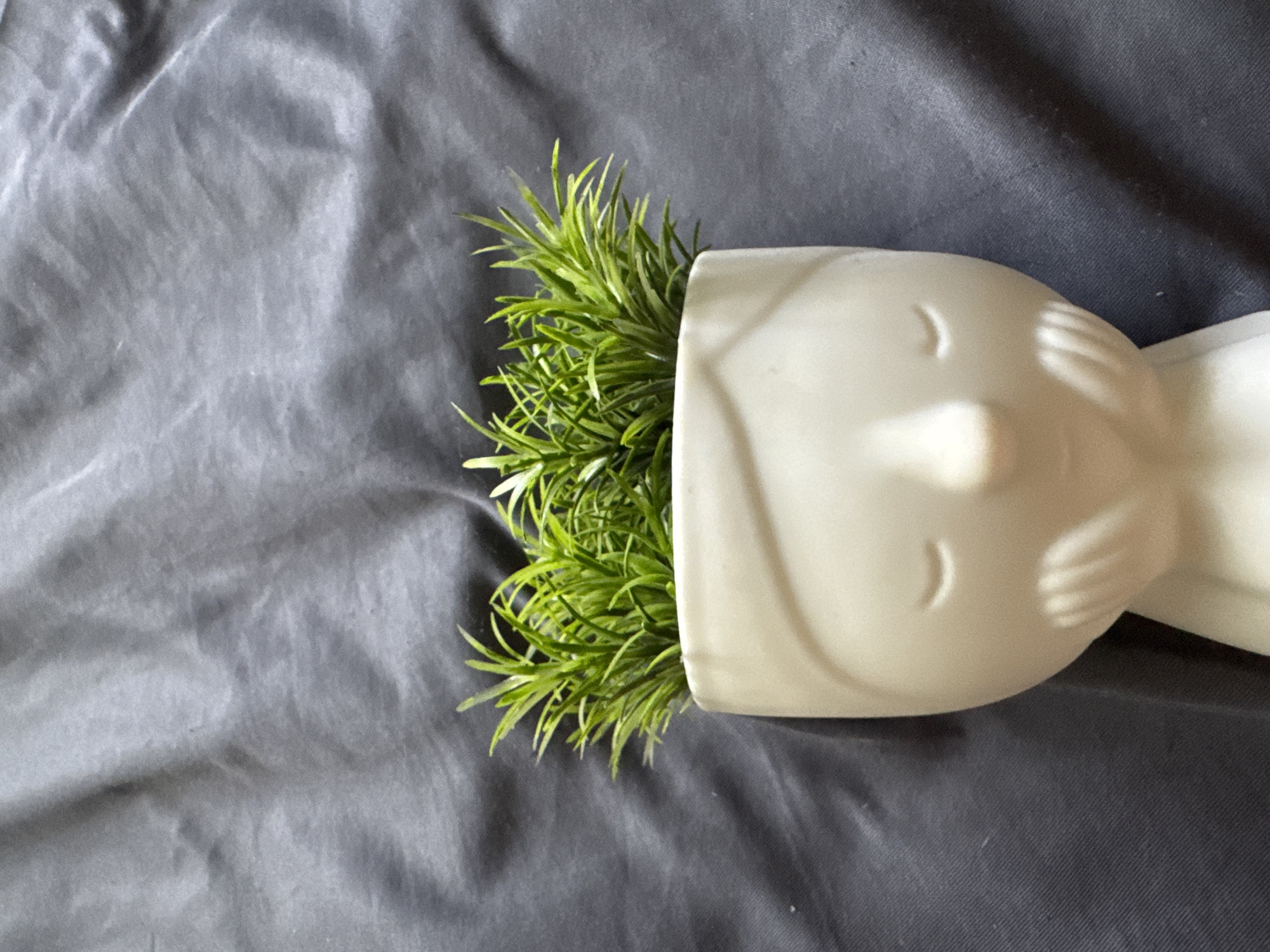 A close-up, top-down shot features a white ceramic planter shaped like a stylized head with closed eyes, resting on a dark grey, creased fabric surface. Green, grass-like artificial plants sprout from the top of the planter. The scene is photographed indoors, with no specific location in Sofia, Bulgaria, indicated by the visual elements.