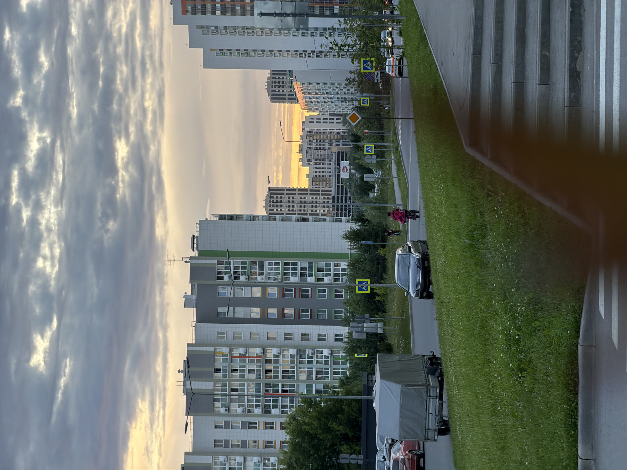 The image depicts a street scene in Kazan, Russia, during what appears to be either dawn or dusk. Tall, modern residential buildings line one side of a road, with a strip of vibrant green grass separating the buildings from the street. The sky is filled with dramatic, streaky clouds, illuminated by the soft glow of the setting or rising sun, suggesting a dramatic weather event or time of day.
On the road, several cars are visible, including a dark sedan and a truck with a covered trailer. Pedestrian crossing signs are strategically placed along the sidewalk, indicating a safe passage for pedestrians. In the middle ground, a person on a bicycle, wearing a red and black plaid jacket, is visible, adding a touch of human activity to the urban landscape. The overall mood is serene, with the soft light and quiet atmosphere of a residential area. The buildings themselves are multi-story structures with numerous windows, characteristic of urban housing developments. The architecture suggests a contemporary setting, and the presence of Russian street signs would confirm the location as Russia.