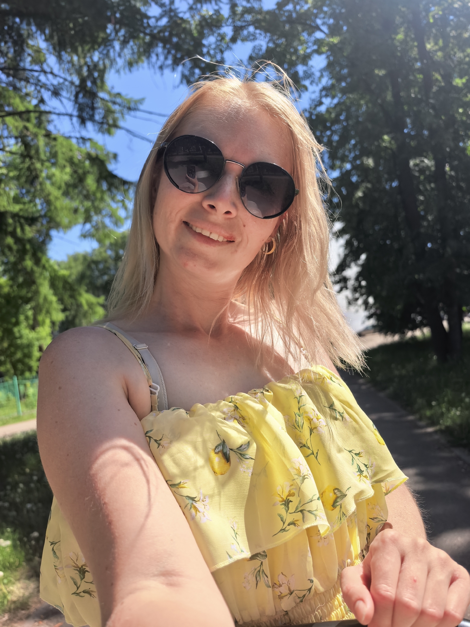 A blonde woman with sunglasses is taking a selfie in a park. She is wearing a yellow floral top. The background shows green trees and a blue sky. There is also a visible road. There is no information to confirm the location in the city of Sterlitamak, Russia.