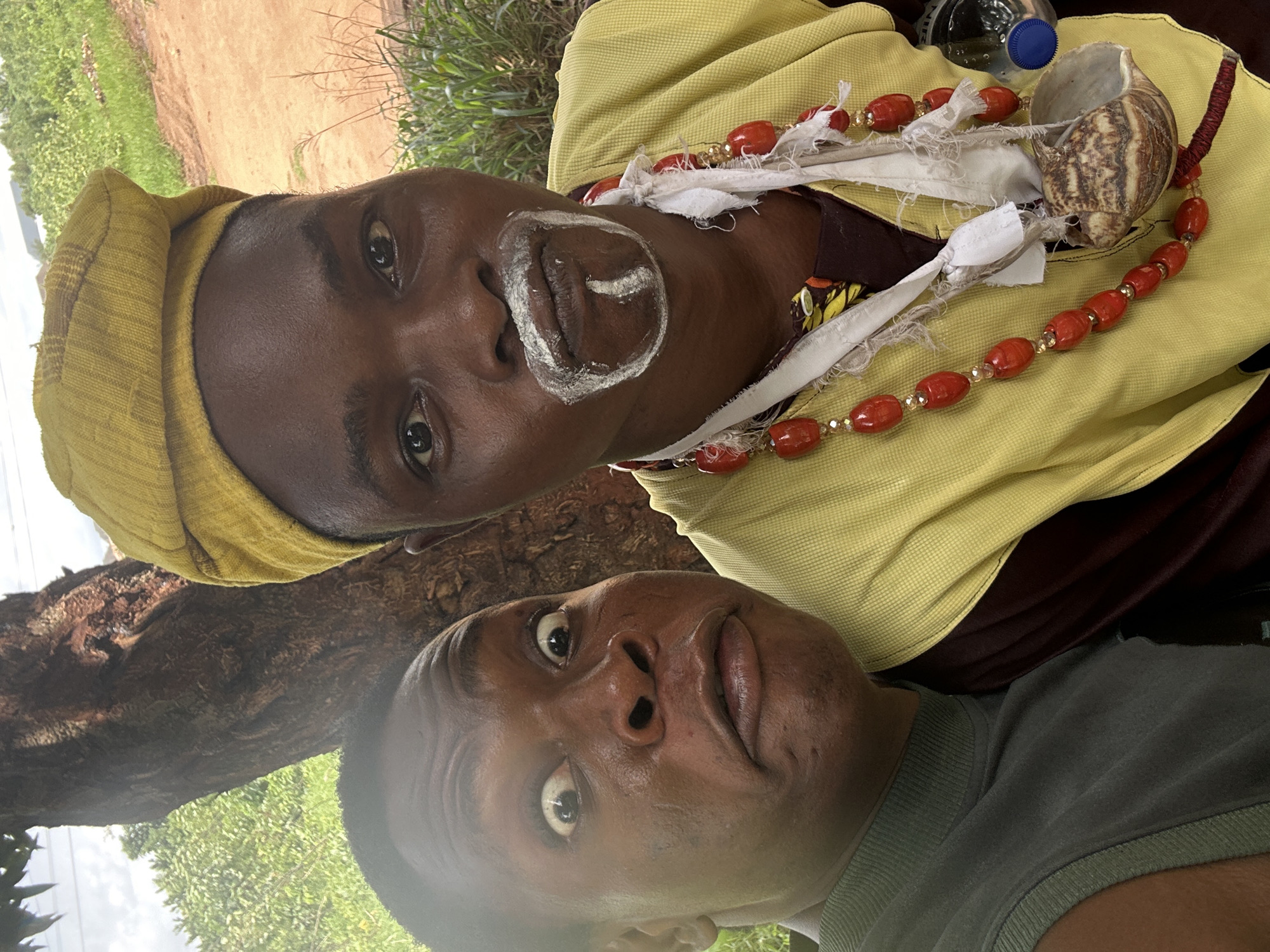 The image depicts two people, likely engaged in a selfie. The individual at the top appears to be wearing traditional attire, including a yellow head covering, a necklace of red beads, and white paint around their mouth. They also have a large snail shell and a water bottle nearby. The person at the bottom is wearing a gray t-shirt. The background suggests an outdoor setting with trees and foliage. The location is Unknown, Unknown.