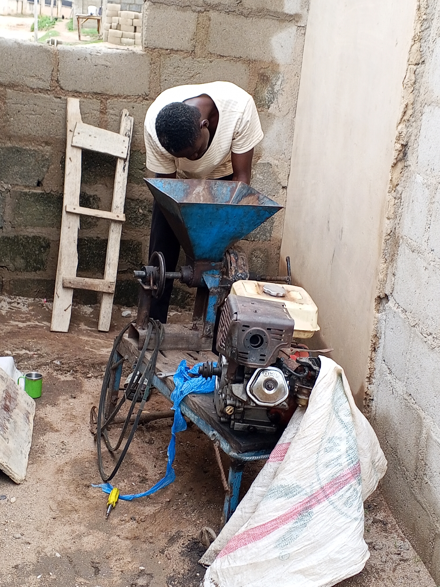 In Zango-ndaji, Nigeria, an individual stands next to a grain milling machine outdoors. The machine is composed of a hopper, grinder, gas motor, and drive wheels. The individual is positioned, leaning over the machine at the hopper. A wooden ladder leans against a grey block wall to the left of the machine. An open-ended green mug sits in front of the wall. A yellow handled screwdriver lies on the ground. A white sack is located near the base of the machine.