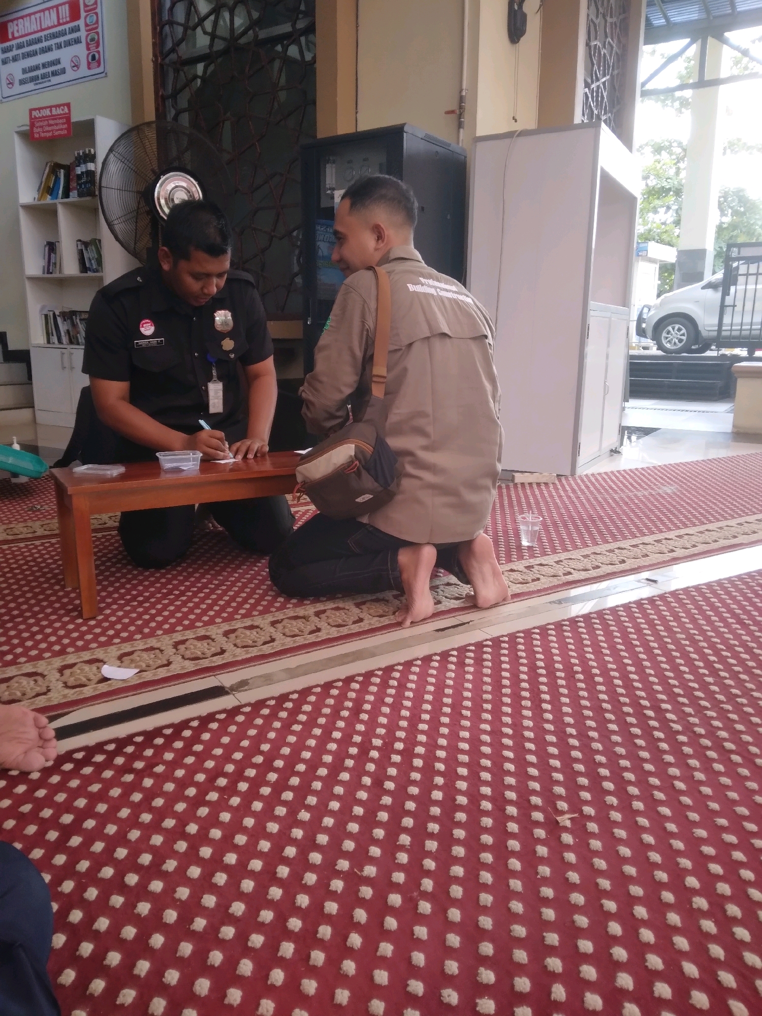 In Desa Pamijen, Kecamatan Sokaraja, Indonesia, two men are engaged in an activity indoors on a red carpeted floor. The man on the left, wearing a black uniform with badges and a name tag that appears to read "MAMUARI Y." is kneeling on the floor. He is facing a small wooden table and is in the process of writing on a piece of white paper with a blue pen. Next to the paper is a small clear plastic container. The man on the right, wearing a beige uniform shirt with a bag slung across his shoulder, is also kneeling. He is facing the man in the black uniform and appears to be observing him. In the background, a large industrial fan is visible, along with shelves filled with books. A white cabinet or structure is also present. The environment suggests an indoor public space, possibly a community center or religious building given the carpet pattern. Outside, through a large window or opening, a white vehicle is partially visible.