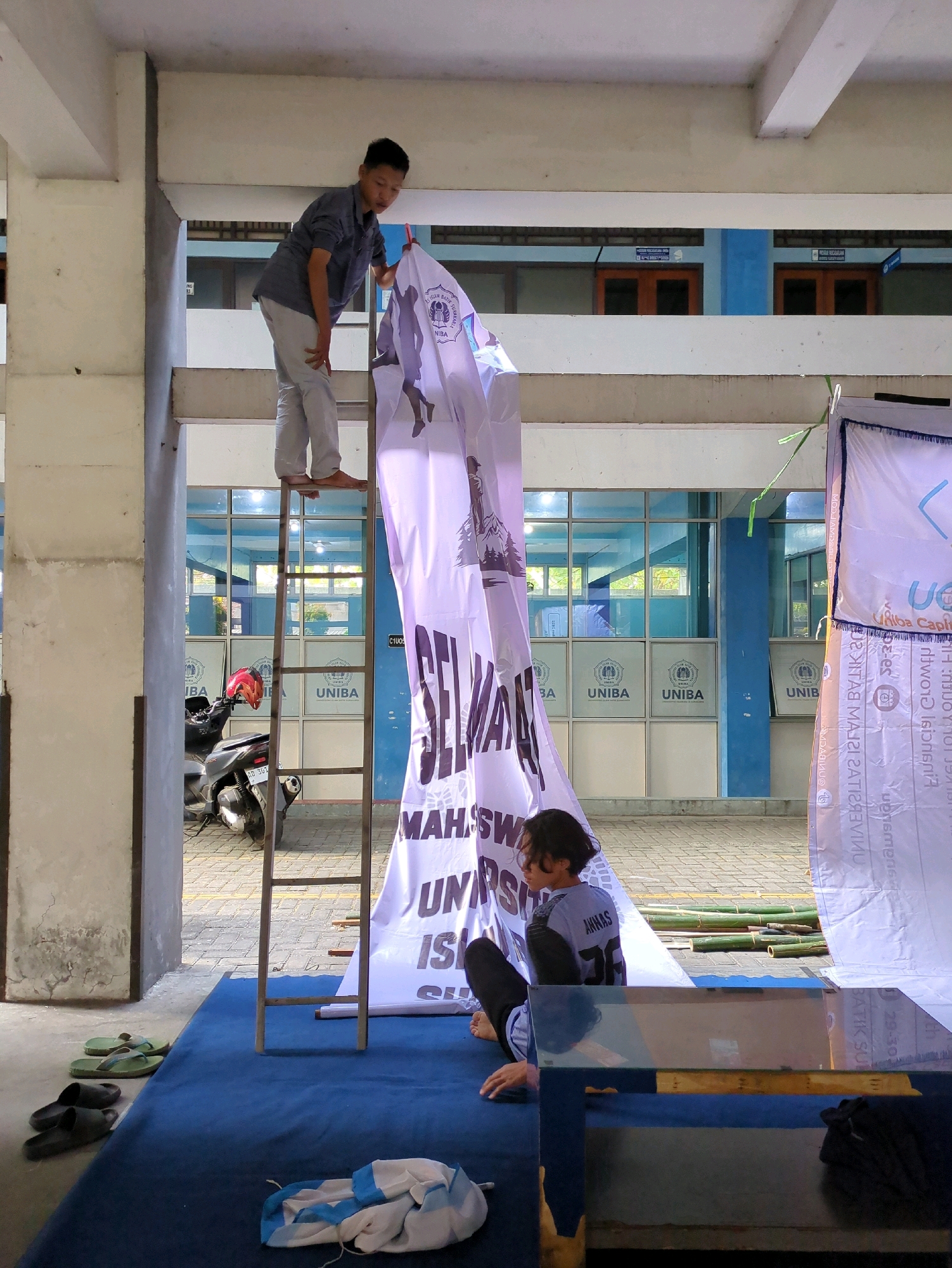In Surakarta, Indonesia, a young man stands on a tall ladder, holding a large white banner with black text and graphics. He is wearing a dark blue collared shirt and light gray pants. The banner is partially unfurled and displays text that appears to be in Indonesian, with some images of people and a logo. Below him, another young man sits on the ground, wearing a dark long-sleeved shirt with "AKNAS 36" written on the back and dark pants. He is looking to his left, with his right arm resting on his knee. A motorcycle with a red helmet is parked to the left of the ladder, visible through a large opening in a concrete pillar. The background shows a building with blue trim and glass windows, revealing multiple signs with logos and text. The ground is paved with brick-like material, and a blue tarp is laid out in front of the banner, with several pairs of flip-flops and a bundled piece of clothing on it. Bamboo poles are visible on the ground behind the banner. The scene appears to be an outdoor setup, possibly for an event or promotional activity.
