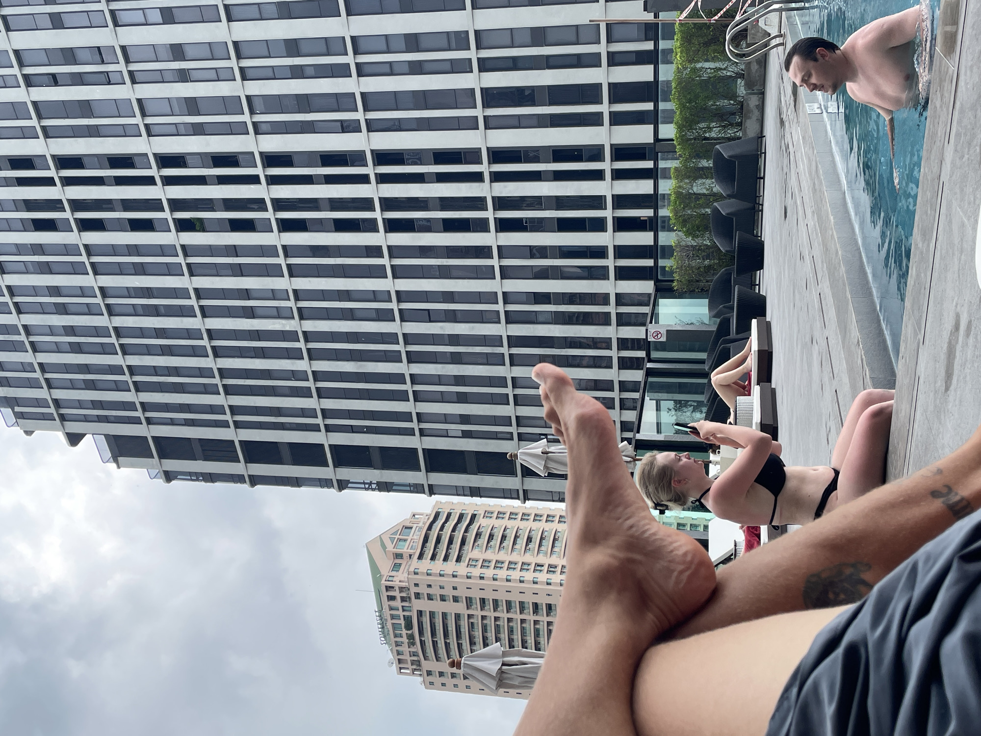 The image, rotated 90 degrees counter-clockwise, offers a first-person perspective from someone relaxing by a swimming pool in a high-rise urban environment. In the immediate foreground, the bare feet and lower legs of the photographer are visible, wearing dark shorts, with a tattoo on the right leg.
To the right, a blonde woman in a black bikini lies on her stomach near the pool's edge, looking at her mobile phone. Further along, a shirtless man with dark hair is partially submerged in the blue pool water, looking downwards. The pool area features concrete decking, dark-colored lounge chairs, and some green bushes. White sun umbrellas are also scattered in the mid-ground. A white sign with a faint 'no smoking' or similar symbol is partially visible near the pool.
The background is dominated by two prominent buildings against an overcast, grey sky. A tall, modern skyscraper fills the left and top of the frame, characterized by its repetitive pattern of dark windows within a stark white or light grey grid-like facade. A small green object, possibly a plant, is visible in one of its windows. Another distinct building, lighter in color (beige/pinkish), is seen further back with multiple balconies, and the word "SOMERSET" is clearly visible in dark letters on its side.
The scene depicts a tranquil daytime activity, suggesting a leisurely moment at a hotel or serviced apartment pool in a bustling city like Bangkok, Thailand, under cloudy skies.