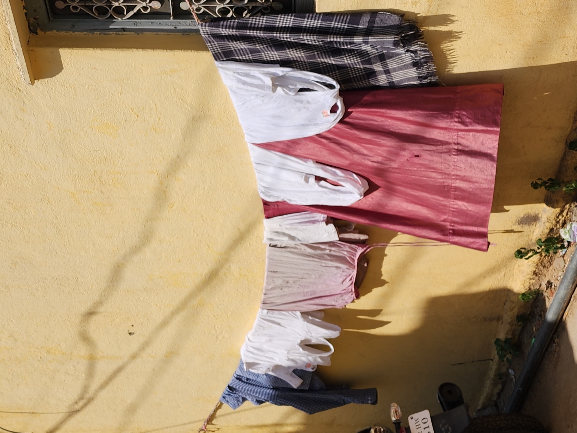 The scene depicts an exterior view of a light yellow stucco wall in Pedda Kammavaripalli, India, under direct sunlight. A rope, functioning as a clothesline, extends horizontally across the central portion of the wall. Seven items of laundry are suspended from this line: a blue and white checkered long-sleeved shirt, three white tank tops, a light purple-pink sleeveless garment, a magenta-red lower garment with a visible drawstring, and a dark plaid shawl with fringed edges. Strong, defined shadows from the clothesline and the garments are cast upon the wall, indicating illumination from the right. On the upper right section of the wall, a window with a dark frame and intricate metal grilles is present. The lower left quadrant shows the rear wheel and a portion of the fender of a black motorcycle or scooter, bearing a white license plate with the registration "AP 32 HW 510". The ground directly beneath the wall is unpaved, featuring small green plants and a dark pipe running horizontally. No individuals are visible.