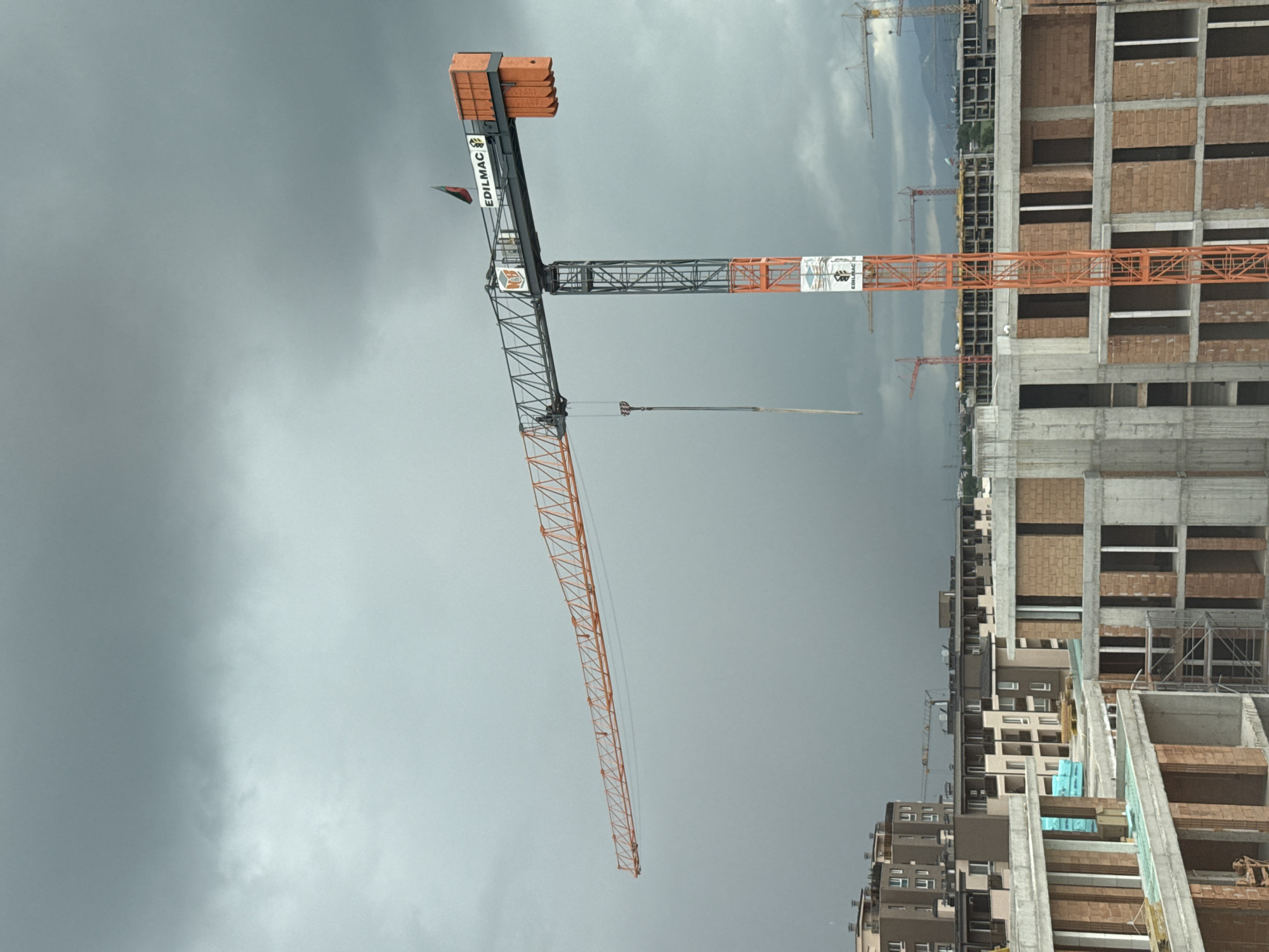 The image depicts a construction site in Plovdiv, Bulgaria, under a heavily overcast sky. A large "EDILMAC" brand tower crane dominates the left side of the frame, its orange boom extending diagonally across the sky. Adjacent to the crane is an unfinished high-rise building with exposed brickwork, concrete supports, and open window spaces. Additional construction cranes are visible in the background, near other buildings. Some of the other buildings appear to be residential.