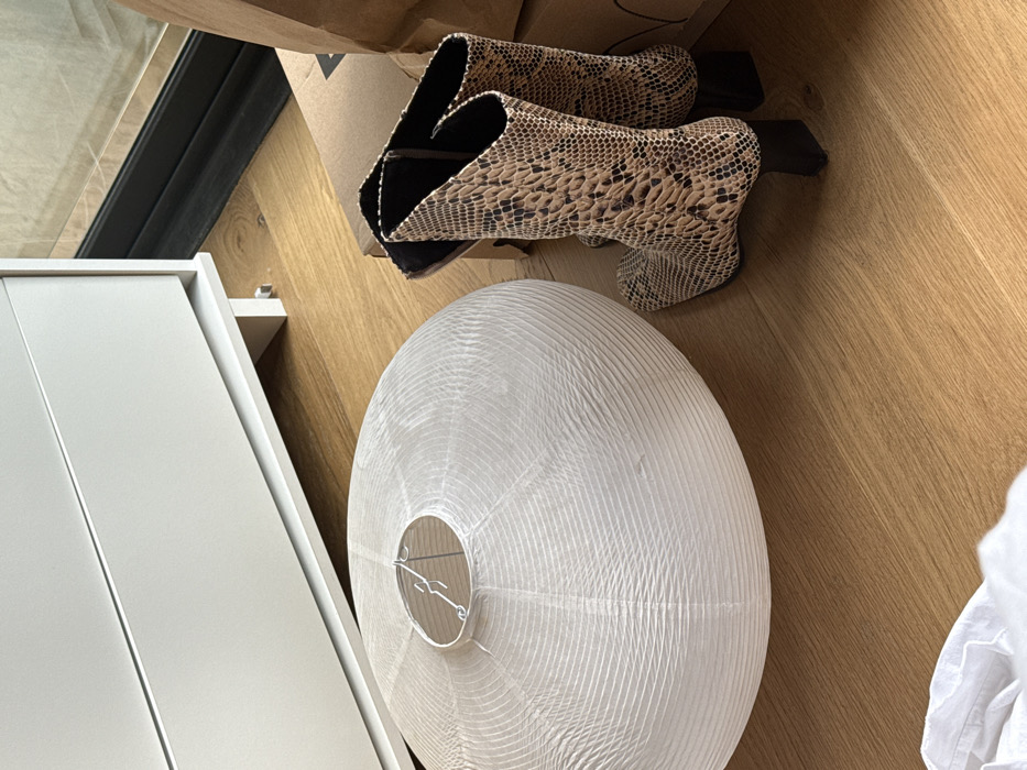 The scene is an interior view, presumed to be in Mexico City, Mexico, depicting household objects placed on a light-colored wooden floor. A large, flattened white paper lantern, approximately spherical in shape with horizontal ribbing and a visible metal internal frame, rests on the floor in the left foreground. To its right, a pair of mid-calf height women's boots with a tan and dark brown snakeskin pattern and black block heels stand upright. In the background, a white dresser or cabinet with two visible drawer fronts is positioned against a wall. Further to the right in the background, two light brown cardboard boxes are partially visible, along with a dark vertical frame, possibly part of a window or door. A crumpled white fabric is partially visible in the bottom-left foreground. No individuals are present.