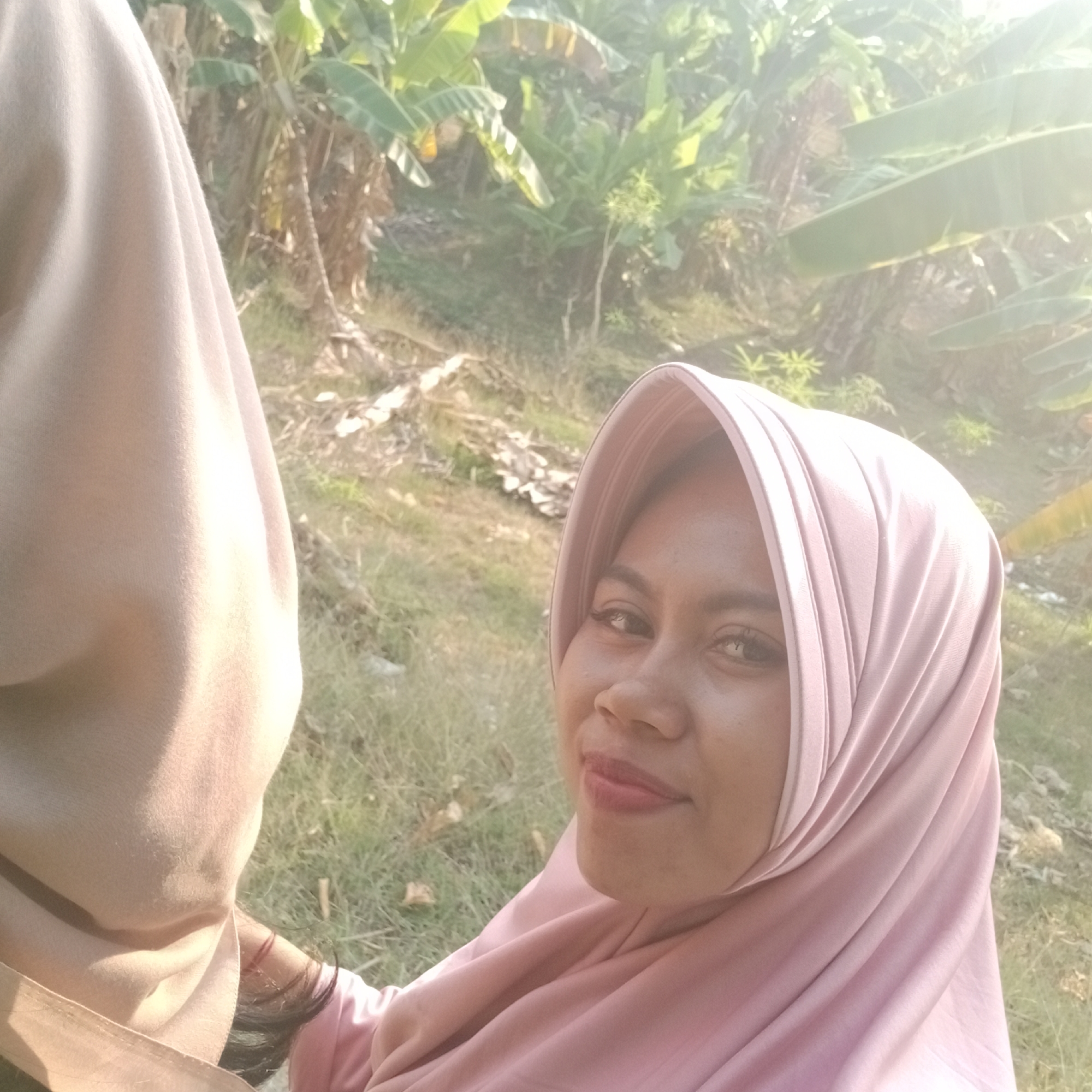 A selfie captures two women from the chest up, posed in a landscape. The woman on the right, wearing a pink hijab and a pink top, is looking at the camera with a slight smile. She has dark eyes and light brown skin. The woman on the left is partially visible, her face obscured by a beige garment, possibly another hijab or scarf. Her skin tone is similar to the woman on the right. The background features lush vegetation, including large banana leaves, indicating a tropical or subtropical environment. Sunlight filters through the leaves, creating a bright, slightly overexposed scene with visible lens flare. The ground is covered in dry grass and some scattered debris. The image appears to have been taken outdoors, likely in a rural area. The location is specified as Tegowanu Kulon, Indonesia, which aligns with the tropical vegetation and possibly the subjects' attire.