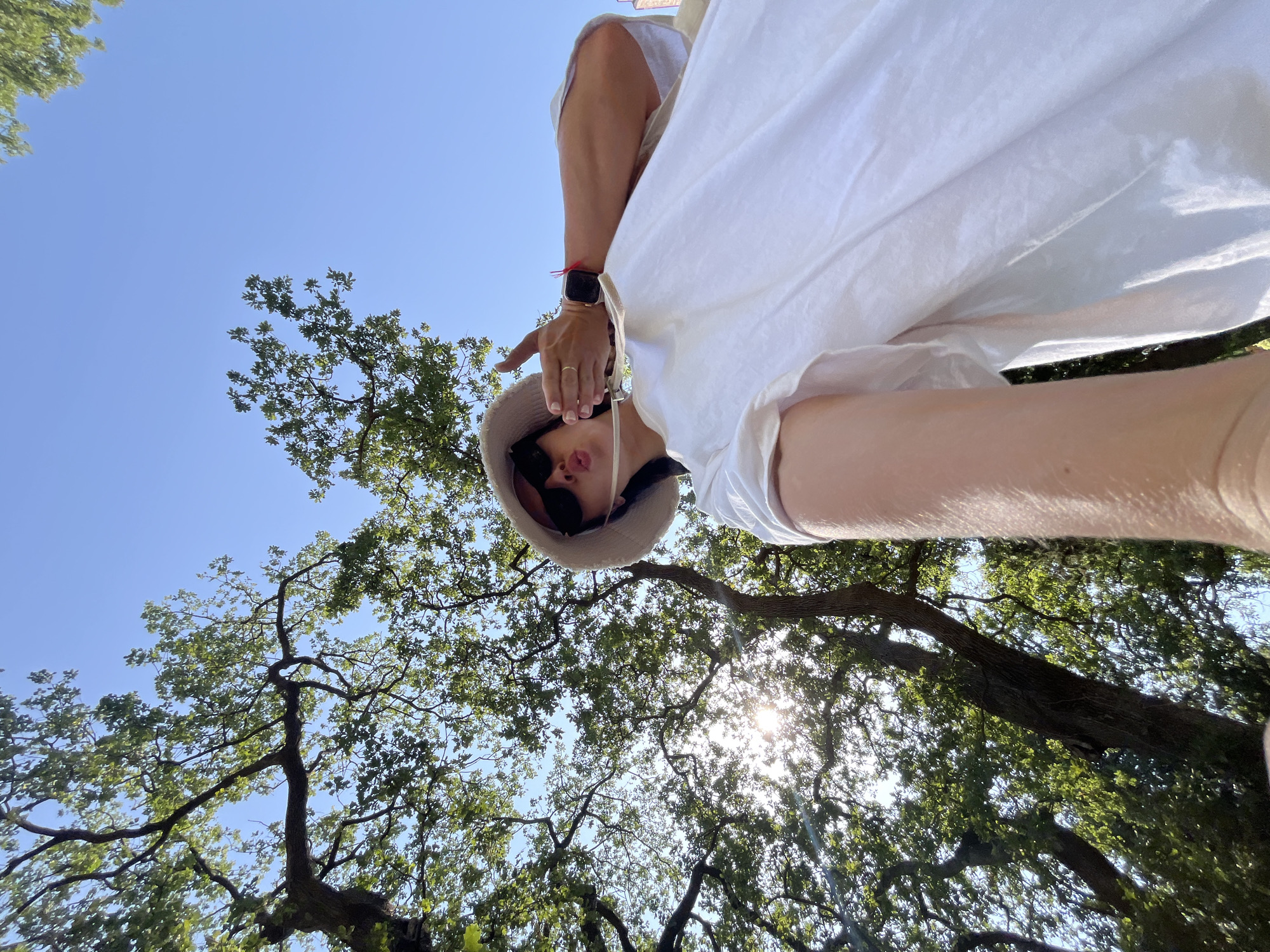 An outdoor, low-angle, eye-level shot captures a young woman in a white oversized t-shirt and a light-colored bucket hat, posing for a selfie under a canopy of green trees. Her lips are pursed, and she is holding up a hand in a peace sign. The sun shines through the trees, creating a lens flare. A smartwatch is visible on her left wrist. The background consists of a clear blue sky and the branches and leaves of trees, suggesting a park or wooded area. The location is identified as Boreti, Montenegro.