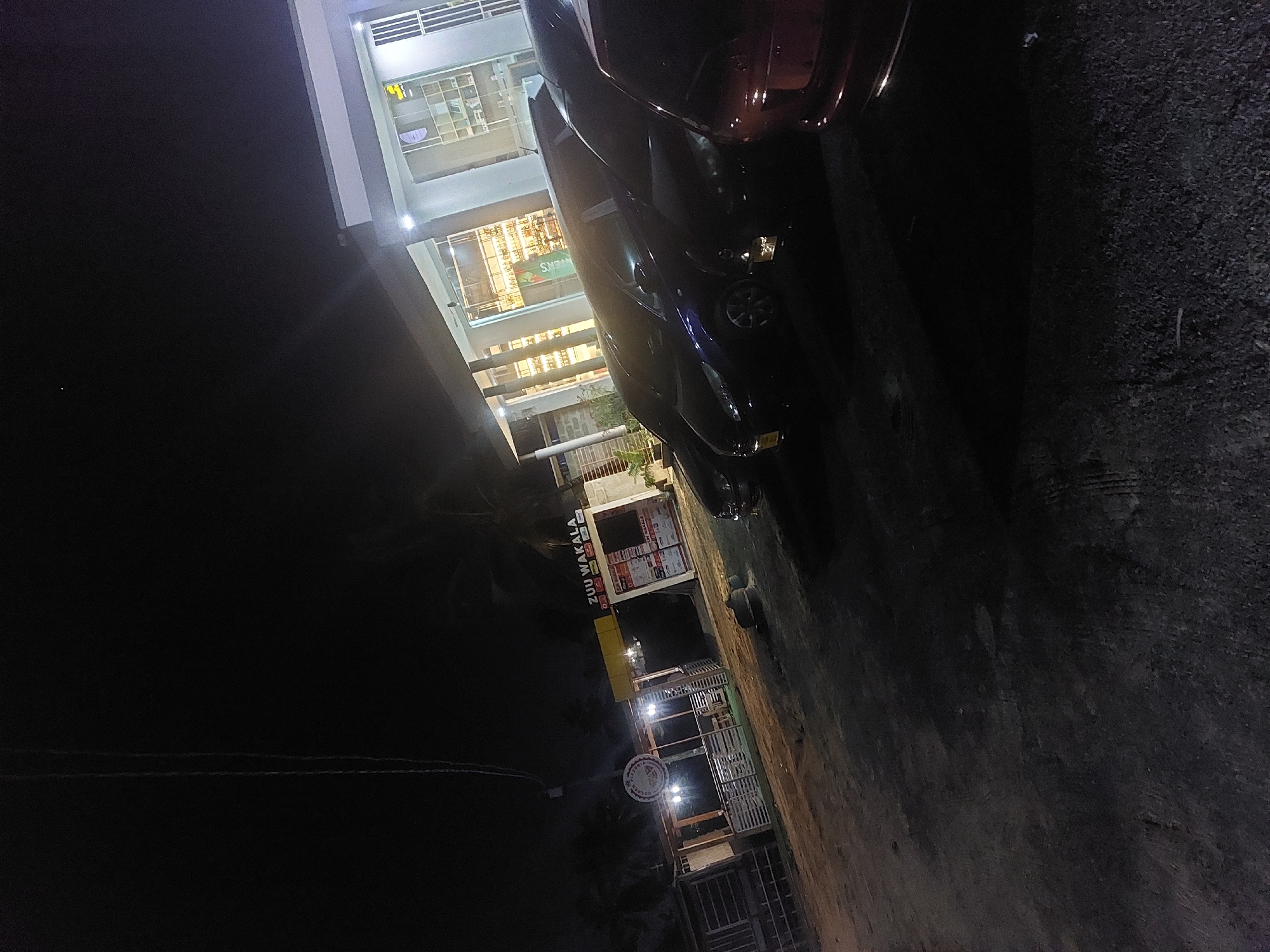 The image depicts a nighttime scene on a street in Dar es-Salaam, Tanzania. In the background, a multi-story building with brightly lit storefronts is visible. A dark-colored car is parked at the side of the street near the building. Adjacent to the road is a local business called "Zuu Wakala" with a yellow sign. A seating area of this business has a partially visible round sign lit up with illumination from the business. The street's surface appears to be paved and dark.