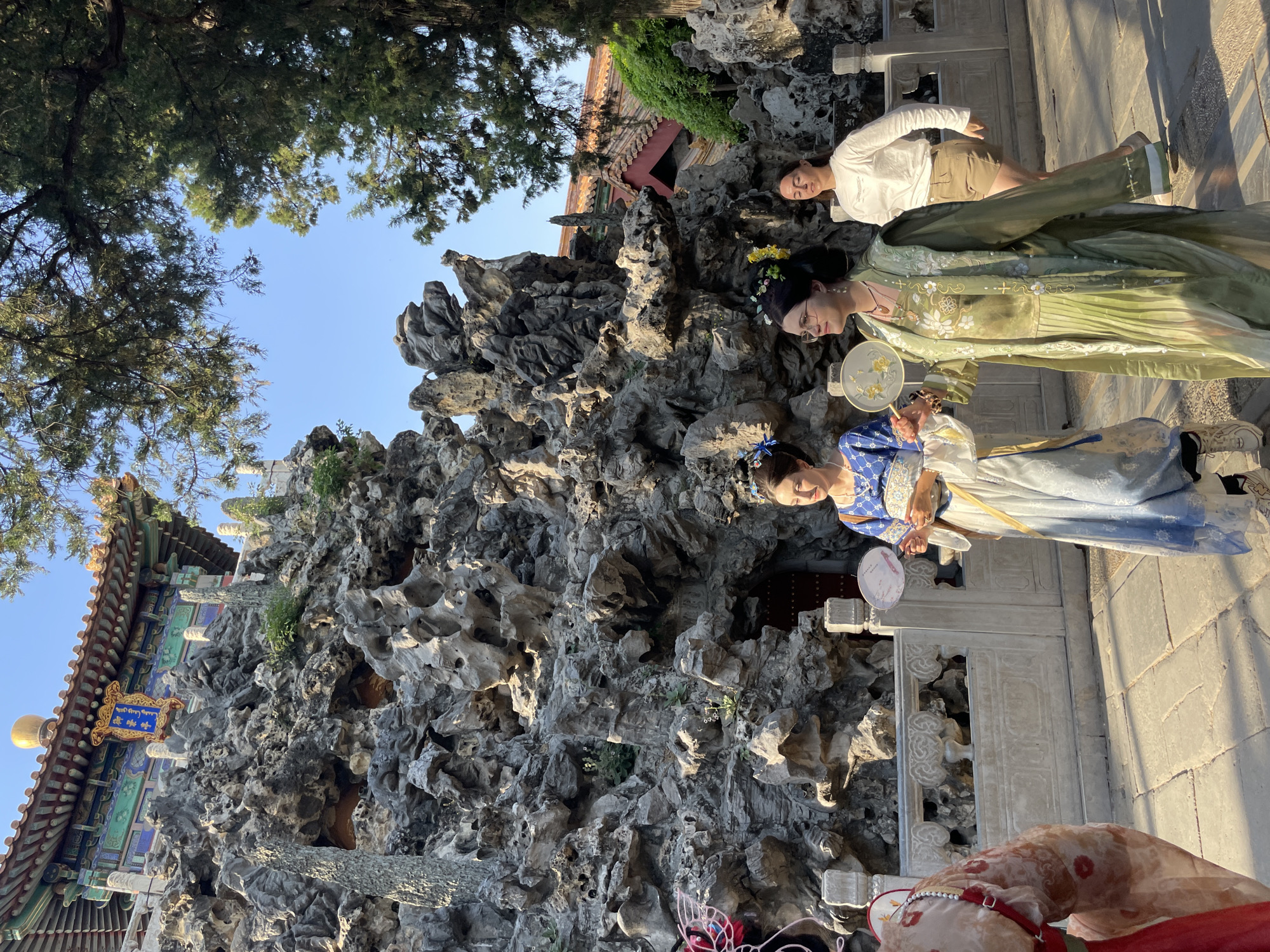 The image depicts a group of three people posing for a photo in front of a rock formation and traditional Chinese architecture.
Two of the individuals, both women, are wearing traditional Chinese clothing, likely Hanfu. One is in shades of blue, holding a fan, while the other wears green. The third person is wearing modern clothing. They are standing on a stone pathway next to the rock formation. The background includes parts of a traditional building with a curved roof and ornate decorations. There are trees overhead and to the side. The location is within the Dongcheng District, China, suggested by the architecture and attire.