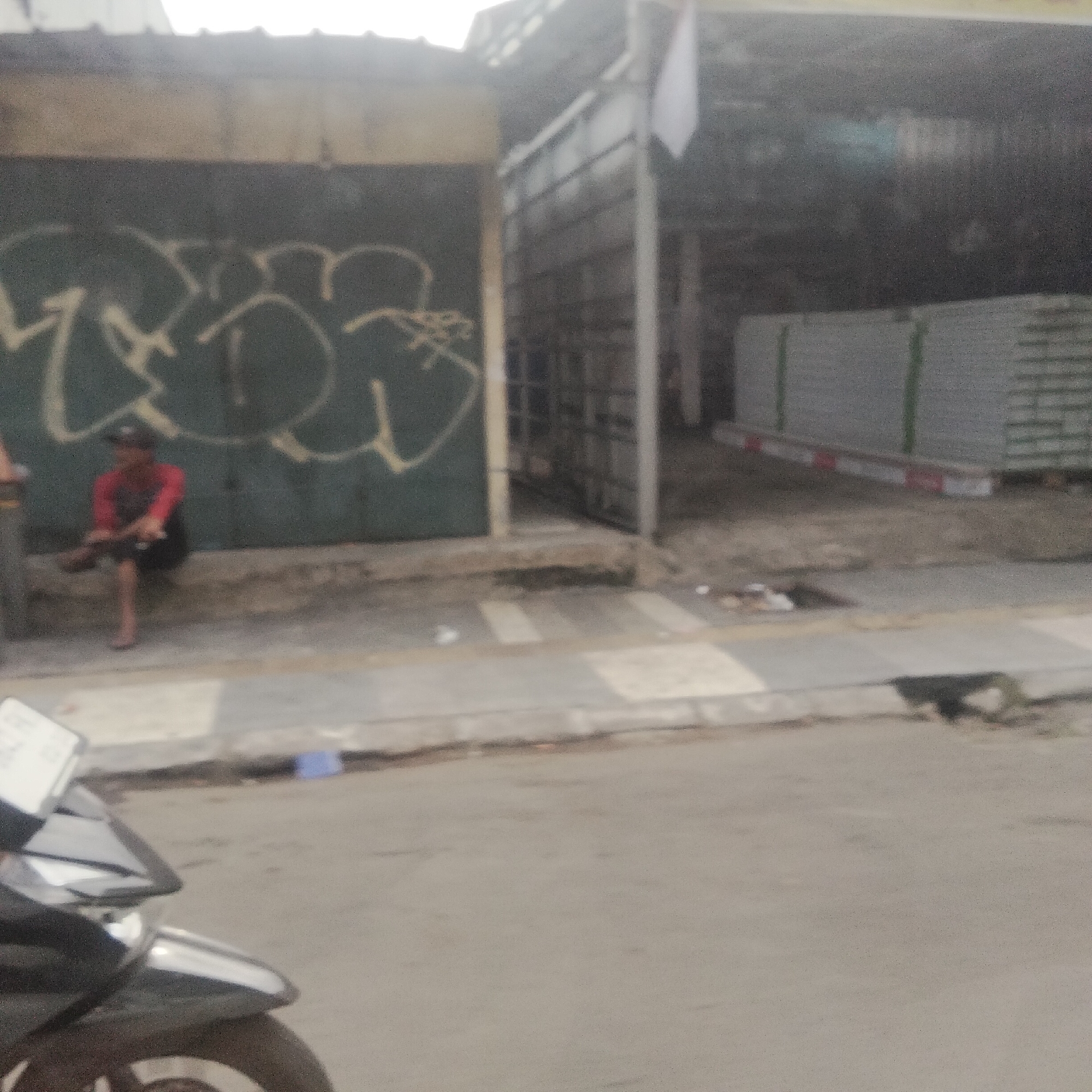 The image depicts a street scene in Bogor, Indonesia, with a focus on an outdoor area adjacent to a building. In the foreground, a portion of a parked motorcycle is visible. The middle ground features a paved sidewalk with a person sitting on a low wall in front of a dark-colored wall covered in graffiti. The person is wearing a red long-sleeved shirt and dark shorts, and appears to be looking down. Behind the seated individual, the building has a section with metal bars and a roof. Further in the background, stacks of what appear to be building materials, possibly plasterboards, are neatly arranged. The overall scene is slightly out of focus, with a grainy texture, and the lighting suggests it is daytime, possibly overcast. There is some debris on the ground near the building.
