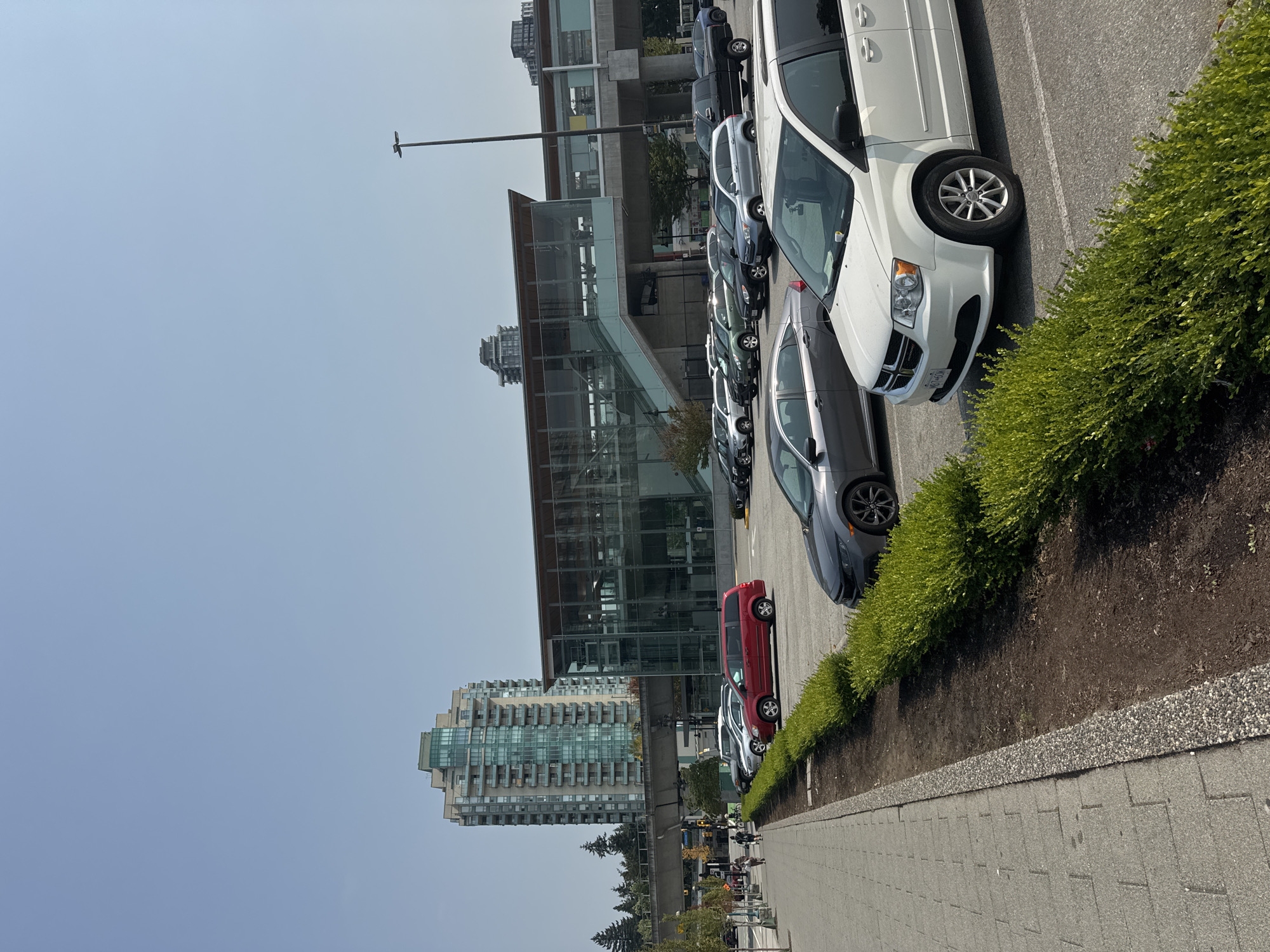 The image shows a parking lot adjacent to a modern building with large glass windows and elevated walkways, likely a public transit station in Coquitlam, Canada. A tall, light-colored apartment building with many balconies is visible in the background, along with trees and a clear sky. Several cars are parked in the lot, including a white minivan in the foreground. A pedestrian walkway with a textured surface runs along the left side of the frame, bordered by a manicured hedge. A few people are walking on the sidewalk. The scene appears to be a sunny day.