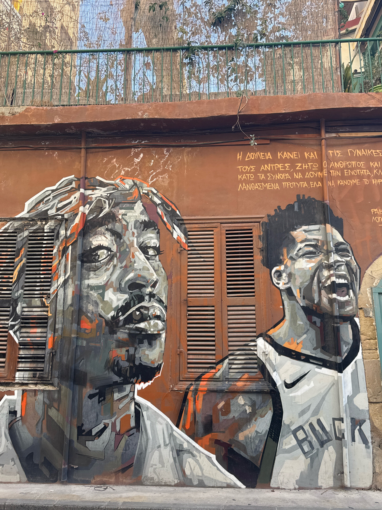 The image depicts a large mural on a building in Limassol, Cyprus. The mural features two portraits of African American men. The man on the left is depicted in profile, looking upwards, with a bandana on his head. The man on the right is shown from the chest up, with his mouth open in a shout or laugh, wearing a basketball jersey with the word "BUCK" visible. The background of the mural is a reddish-brown wall with wooden shutters on either side of the portraits. Above the mural, there is text written in Greek. The overall style of the mural is expressive and uses a combination of grey, black, orange, and white colors. The building appears to be an older structure, with some wear and tear visible on the wall. The foreground shows a concrete sidewalk.