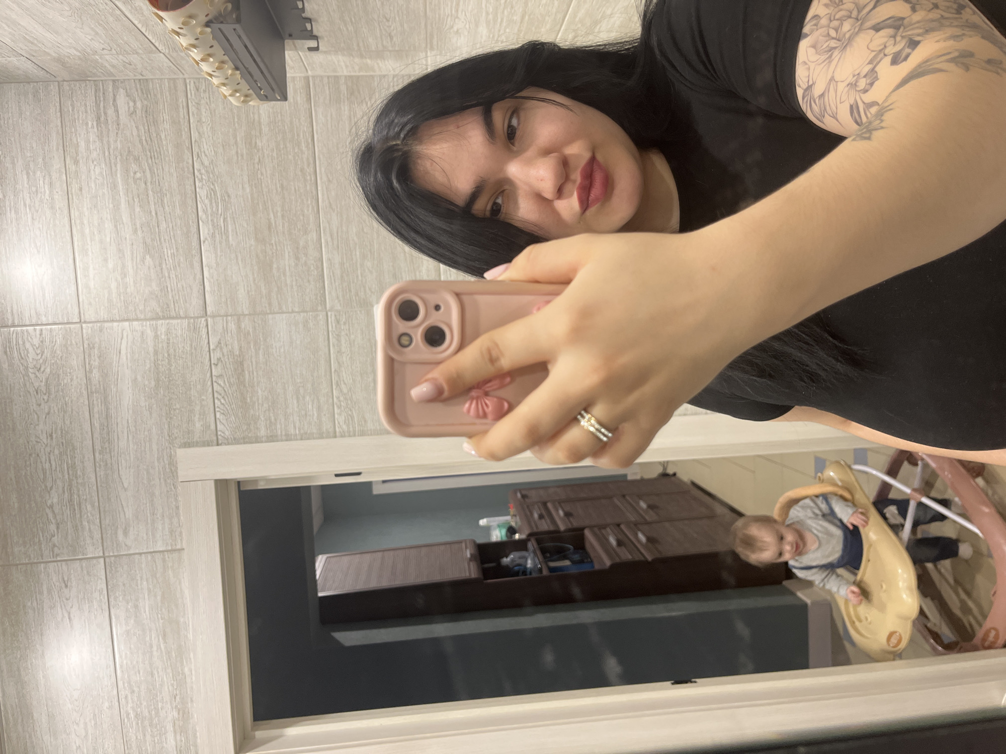 An adult female is captured in a mirror selfie, holding a pink smartphone with a pink bow decoration in her right hand. She has long dark hair, is wearing a black t-shirt, and has a floral tattoo on her left bicep. A two-band ring is visible on her left ring finger. Her lips are slightly pursed. The wall directly behind her is tiled with light-toned, wood-grain patterned rectangular tiles. To the upper right, a dark gray wall-mounted shelf with hooks holds a light-colored, perforated cylindrical object.
The mirror reflects a doorway with light-colored trim, leading to an adjacent room. This reflected room has dark teal-blue walls and a light-colored floor. Inside this room, a tall, narrow, dark brown cabinet with shelves and drawers is visible on the left, alongside a shorter dark brown cabinet. A baby, wearing a grey long-sleeved shirt and dark blue overalls, is positioned in a light brown baby walker within this reflected room, facing towards the mirror.
The scene depicts an interior residential setting. No specific identifying features are present within the image to determine an exact location within the city of Chaikovsky, Russia.
