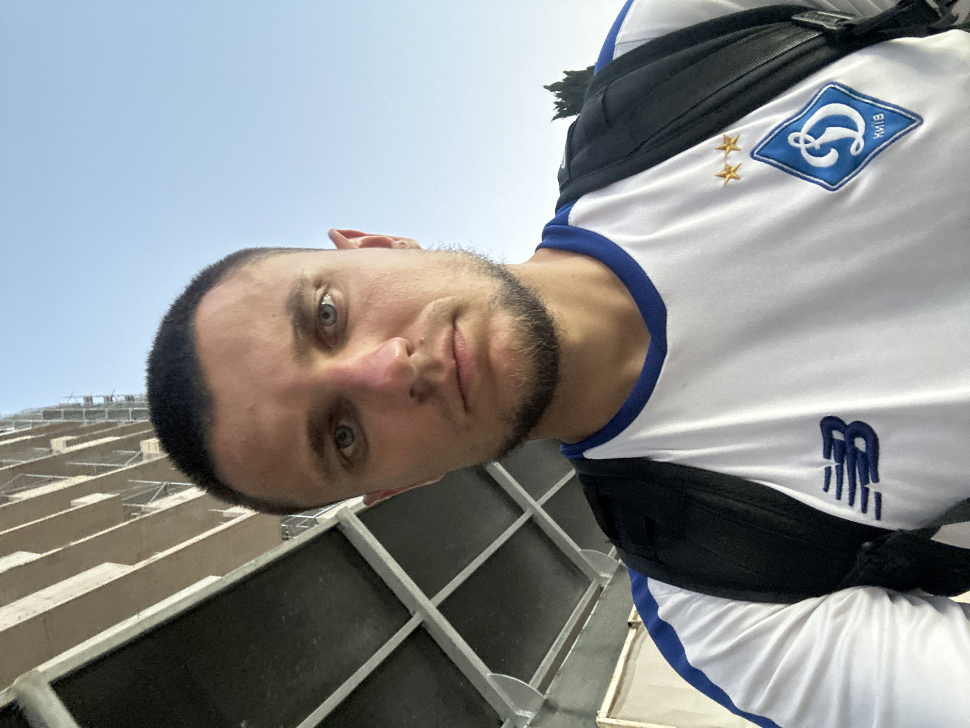 A young adult male is captured in a selfie, looking directly at the camera with a neutral expression. He has short dark hair, stubble on his chin, and light blue eyes. He is wearing a white t-shirt with blue trim around the collar and sleeves. The t-shirt features a blue New Balance logo on the left chest and a blue diamond-shaped crest with a stylized "D" and the word "КИЇВ" (Kyiv) in white, along with two small gold stars, on the right chest. A black backpack is slung over his shoulders. The background shows an unfinished concrete building with exposed rebar and scaffolding, suggesting a construction site. The sky is clear and blue. The overall setting appears to be outdoors, possibly in an urban or semi-urban environment, and based on the provided information, it is identified as being in the city of Budva, Montenegro.