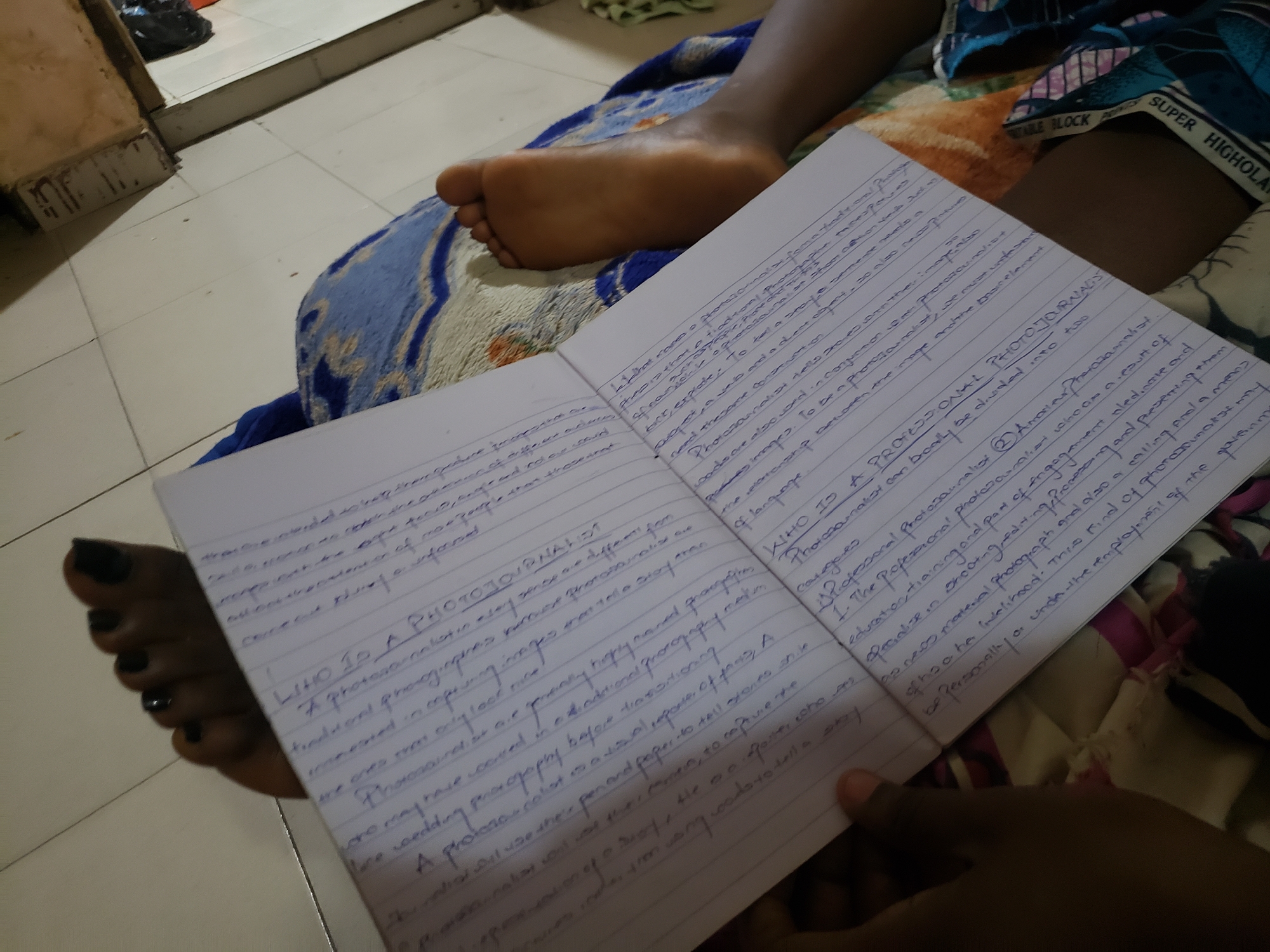 The image displays an indoor scene in Rido, Nigeria, featuring a partial view of an individual. A bare left foot, exhibiting dark nail polish on the toenails, rests on a blue and white patterned blanket or cloth. A left hand, partially visible in the lower right, holds an open, blue-lined notebook. The notebook contains handwritten text in blue ink, with discernible headings such as "WHO IS A PHOTOJOURNALIST?" and related descriptive content. The blanket lies on a light-colored, tiled floor. In the upper left background, a segment of a worn wooden doorframe or wall trim is visible.