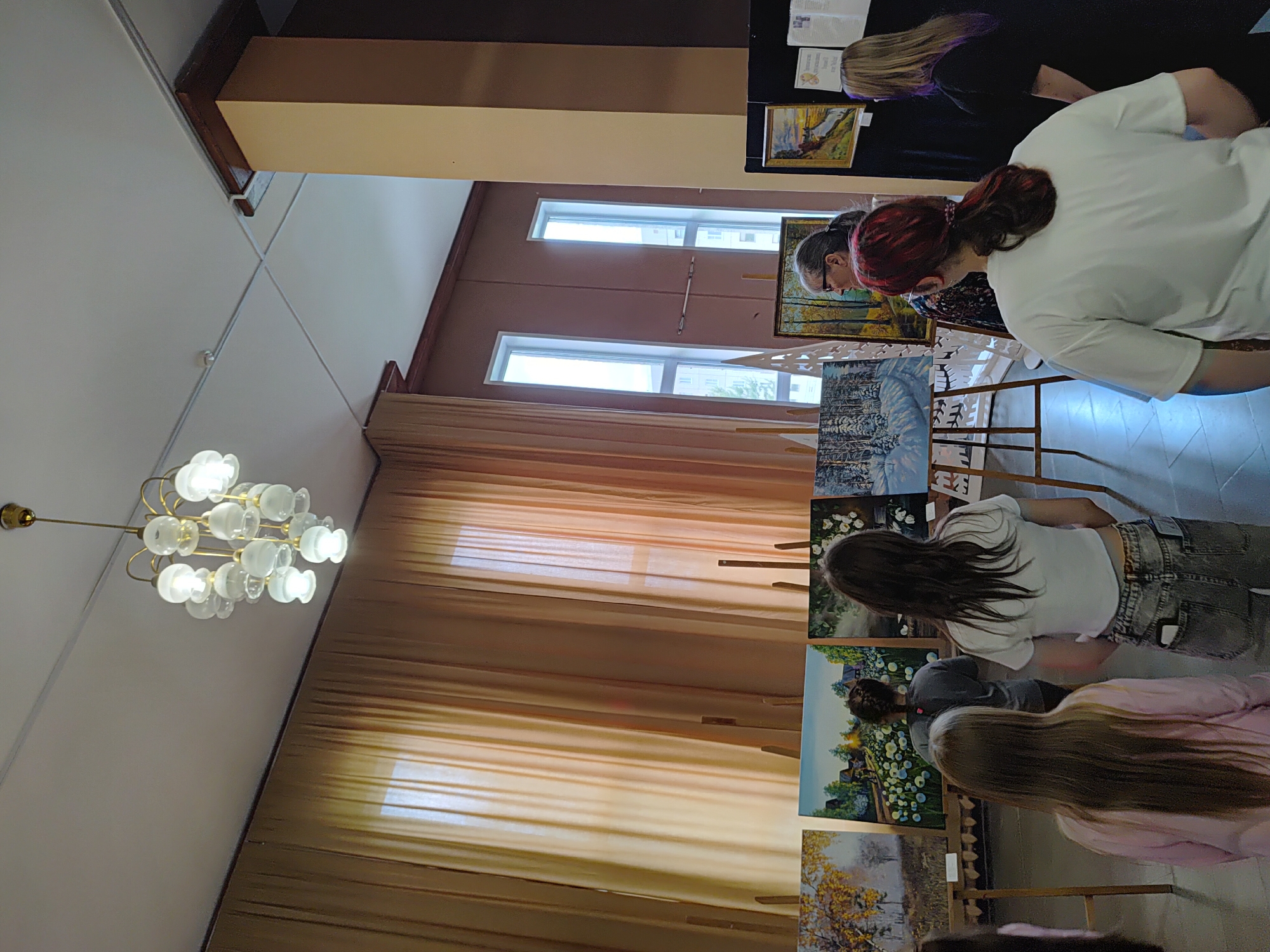 The image depicts an indoor art exhibition, likely in the town of Markovskiy, Russia, given the context. Several people are viewing paintings displayed on easels and a black exhibition board. The room has tall, narrow windows covered by sheer, light-brown curtains, allowing diffused sunlight to enter. A decorative chandelier hangs from the ceiling. The paintings appear to be landscapes, with one in the foreground showing a snowy forest scene and another to its left featuring autumnal trees. In the mid-ground, a woman with glasses and red hair is looking at a painting. In the foreground, three individuals are facing away from the camera, observing the art. One person is wearing a pink shirt, another a white crop top with jeans, and a third a grey t-shirt. The floor is tiled with a dark pattern. The overall atmosphere suggests a cultural event or a school art display.