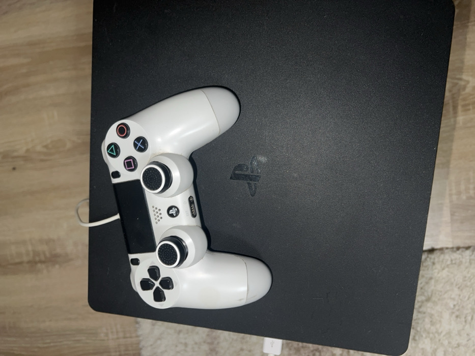 In Sedlartsi, Bulgaria, a white PlayStation 4 DualShock 4 controller rests on a black PlayStation 4 Slim console. A white USB cable connects the top of the controller to the front-left port of the console. The console features an embossed PlayStation logo on its upper surface. The setup is positioned on a light-colored floor, partially covered by a white, textured rug. No individuals are present; the electronic devices are static.