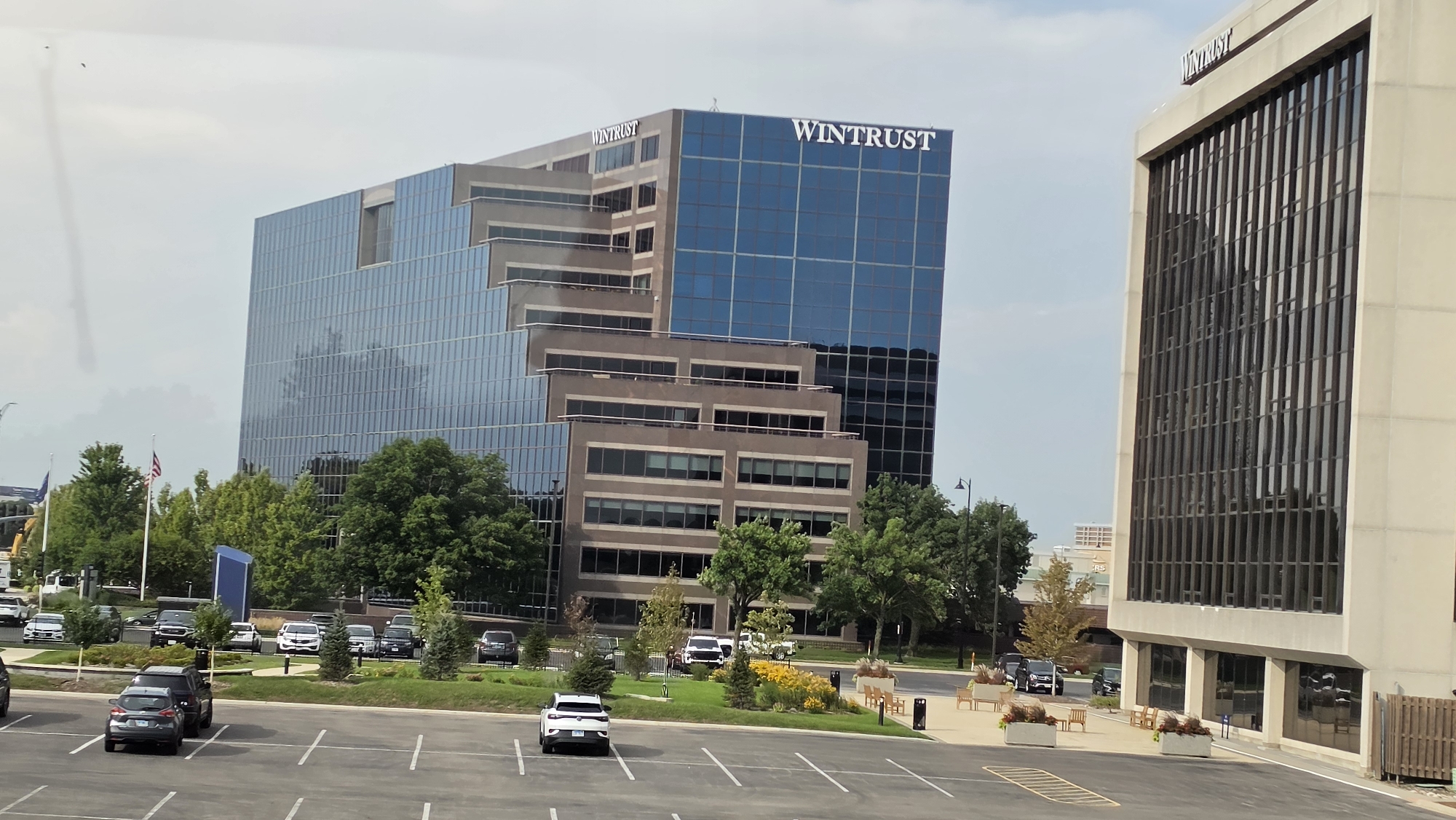 The image shows a view of a modern office building with the "WINTRUST" logo visible on its facade, situated in Rosemont, United States of America. The building is tall and made of glass and concrete, with a stepped architectural design. In the foreground, a parking lot is visible with several cars parked. Surrounding the building are trees and landscaping, including flower beds and benches. To the right of the primary Wintrust building, another office building with a similar architectural style is partially visible. The sky is overcast, suggesting a cloudy day. There are no people clearly visible in the image. The scene appears to be a corporate or commercial district within the city.