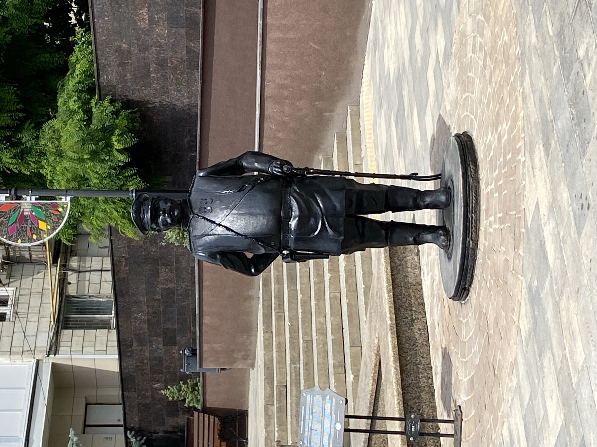 The image shows a full-body statue of a man dressed in a uniform, possibly military or historical, standing on a circular pedestal. The statue is dark in color, appearing to be made of bronze or a similar metal. The man is holding a long object, possibly a sword or a staff, in his right hand, which extends across his body. He is wearing a cap, a long coat, and boots.
The statue is positioned outdoors on a paved area. Behind the statue, there are a set of outdoor stairs leading up to a building with a brown facade. To the left of the statue, there is lush green foliage, and part of a building with a grey tiled exterior is visible. A colorful stained-glass-like decoration is also visible to the far left. The ground around the statue is paved with light-colored rectangular stones laid out in a pattern, with a circular inlay around the pedestal. Sunlight is casting shadows, suggesting it is daytime. The overall scene appears to be a public space, possibly a park, square, or memorial site. There is no visible text in the image.