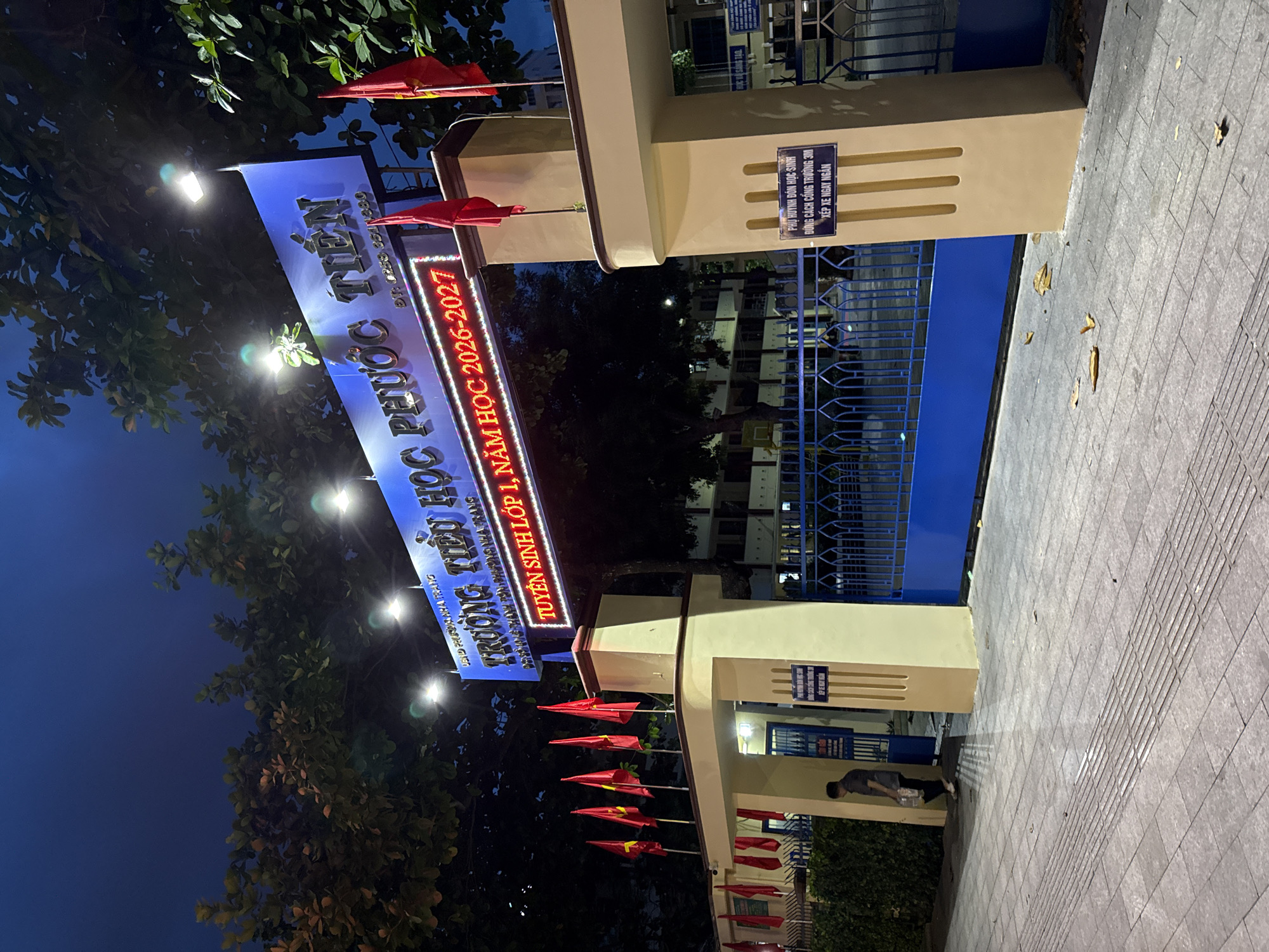 The image depicts the entrance to Truong Tieu Hoc Phuoc Tien, a primary school located in Nha Trang, Vietnam. The school's name is prominently displayed on a blue sign with illuminated lettering. Below the main sign, another illuminated sign reads "TUYỂN SINH LỚP 1, NĂM HỌC 2026-2027," indicating student enrollment for the 2026-2027 academic year. Several red flags with yellow stars, the national flag of Vietnam, are mounted on poles above the entrance.
The entrance itself is flanked by two large, cream-colored pillars. A blue metal gate is visible behind the pillars, with a portion of the school grounds, including buildings and trees, visible beyond. A small blue sign is attached to the right pillar, bearing Vietnamese text related to school rules. A man in a dark shirt and pants is seen walking into the school grounds through an open section of the gate on the left. The scene is illuminated by artificial lights, suggesting it is evening or night. The ground in front of the entrance is paved with grey tiles, and fallen leaves are scattered on the walkway. The sky is dark blue, indicating twilight or night.