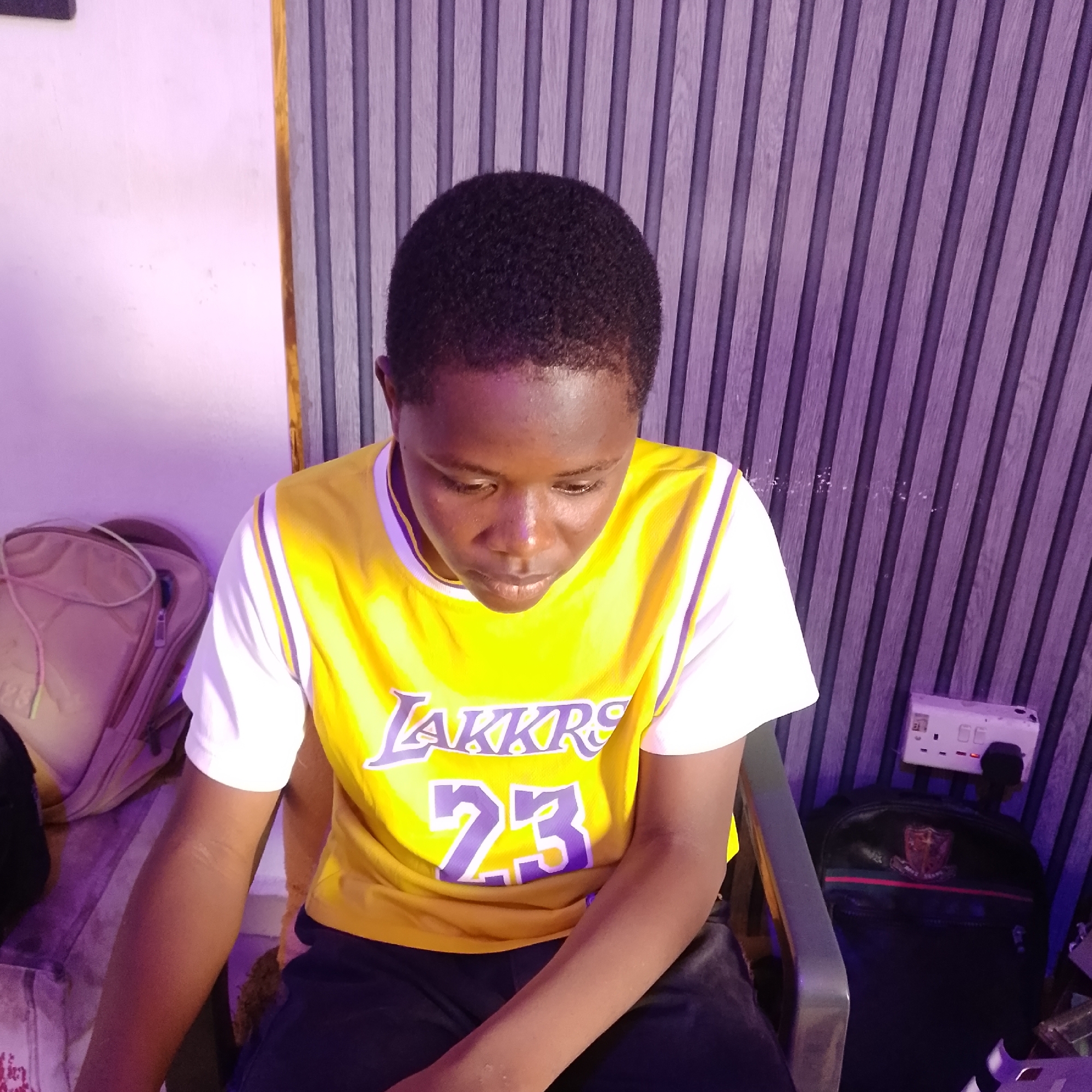 An African youth, wearing a yellow Los Angeles Lakers basketball jersey with the number 23 in purple, sits in a chair in front of a purple-toned, ribbed wall. To the left, a tan backpack rests against a white wall. To the right of the youth, a black backpack is visible next to an electrical outlet mounted on the ribbed wall. The youth is looking down, with their head tilted and eyes focused downwards. The lighting in the scene casts a purple hue.