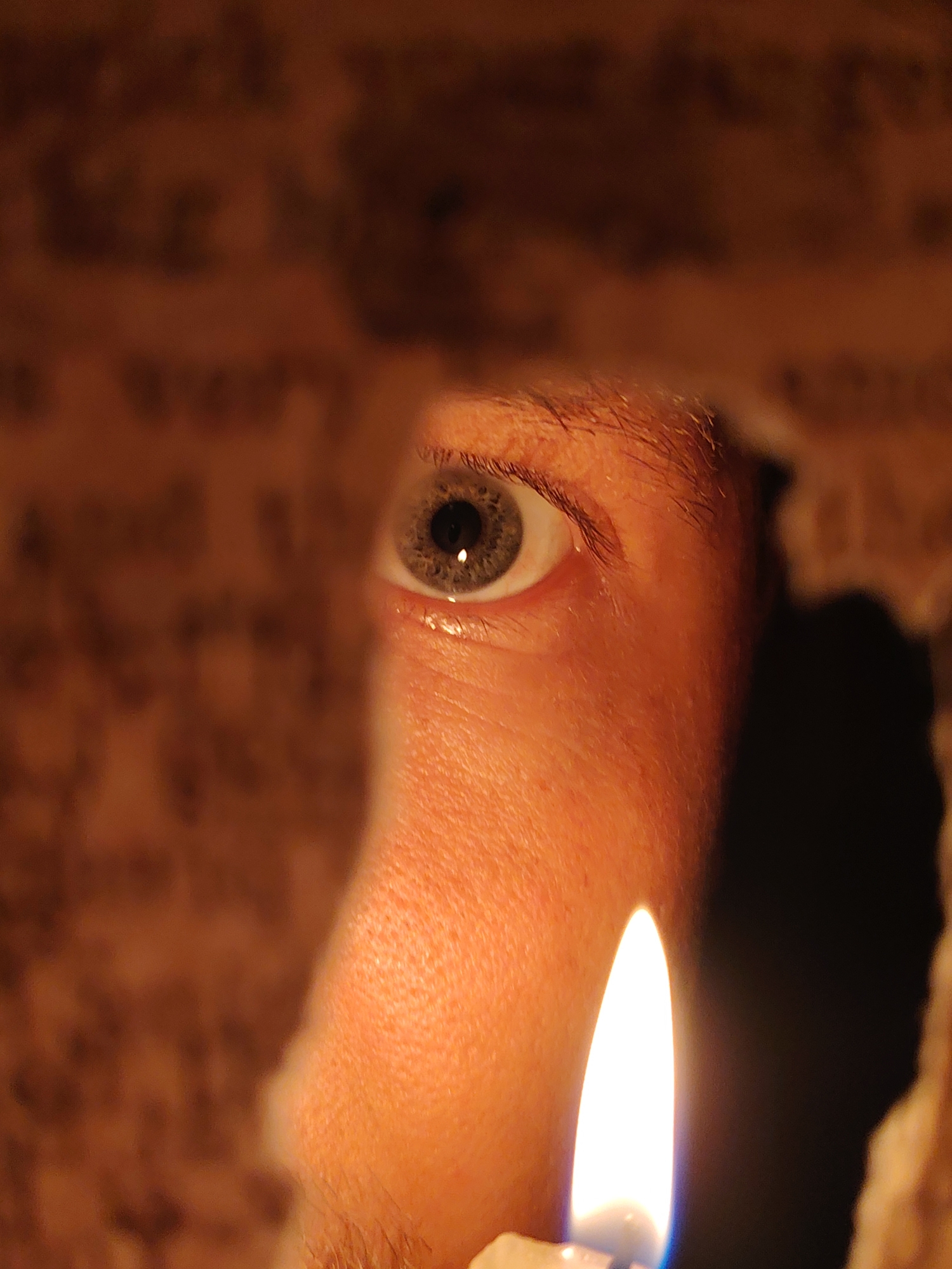 A close-up, eye-level shot focuses on a person's eye peering through a hole in a textured, paper-like material. The eye, with a grey iris and visible eyelashes, is illuminated by a warm, orange light, suggesting a low-light environment, possibly evening or nighttime. The skin around the eye appears flushed, also from the ambient light. To the right of the eye, a bright, white flame from a candle is visible, casting a strong shadow on the right side of the frame and providing the primary light source. The background is a blur of muted browns and oranges, with faint, illegible text appearing on the material the person is looking through. The overall mood is one of intimacy and curiosity, as if the viewer is witnessing a private moment of observation. The textures of the paper-like material and the skin are sharp, contrasting with the soft blur of the background.