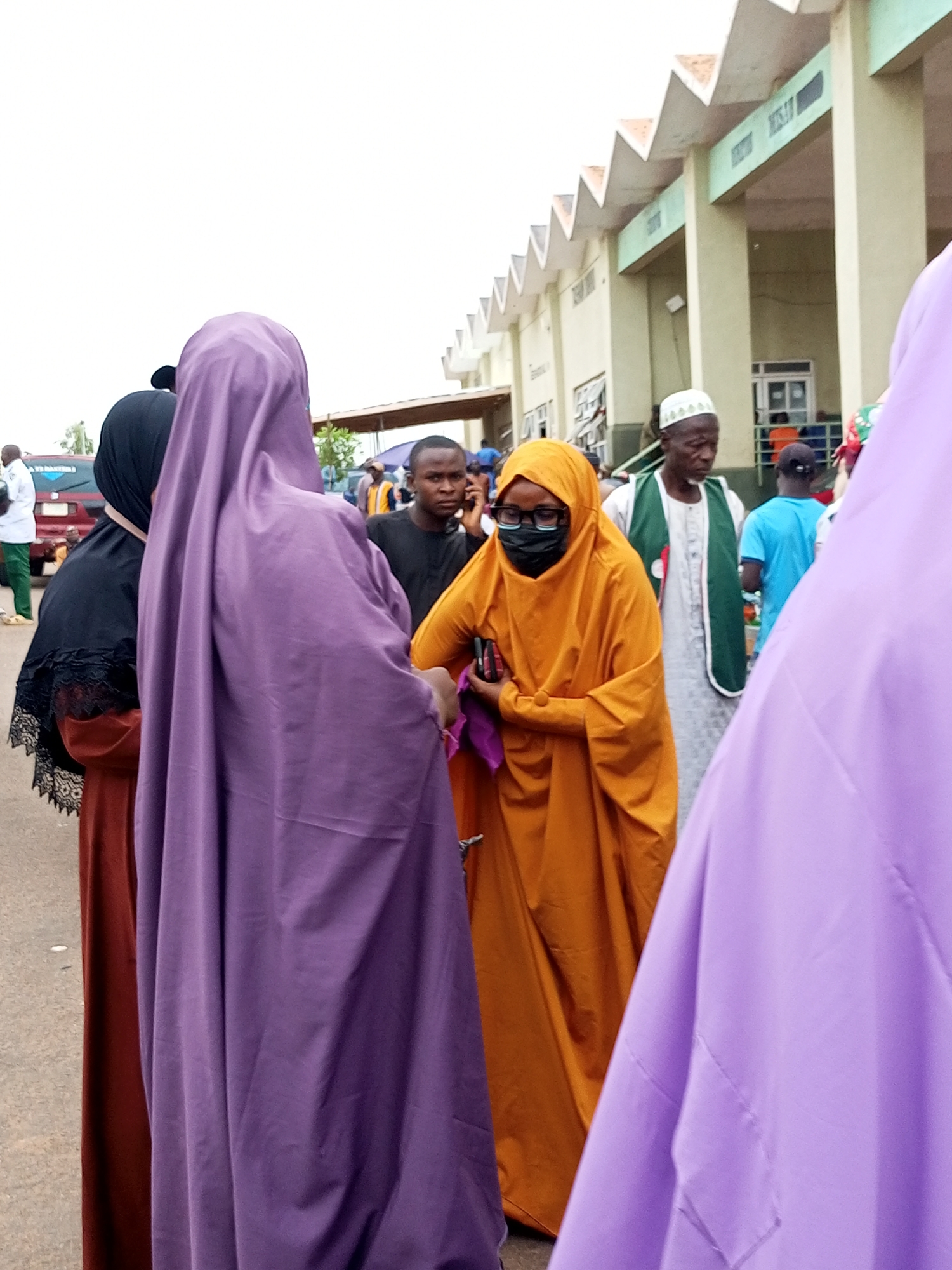 The image depicts a gathering of people outside a building in Gombe, Nigeria. The foreground is dominated by a woman in a purple abaya and another in an orange hijab. Behind them, a man in a black shirt is speaking on his phone, and another man wearing a white cap and green vest is visible. A person in a bright blue shirt is also present in the background. The building behind the people has a distinctive zig-zag roofline and features columns and windows. The overall scene suggests an outdoor event or a busy public space in the city.