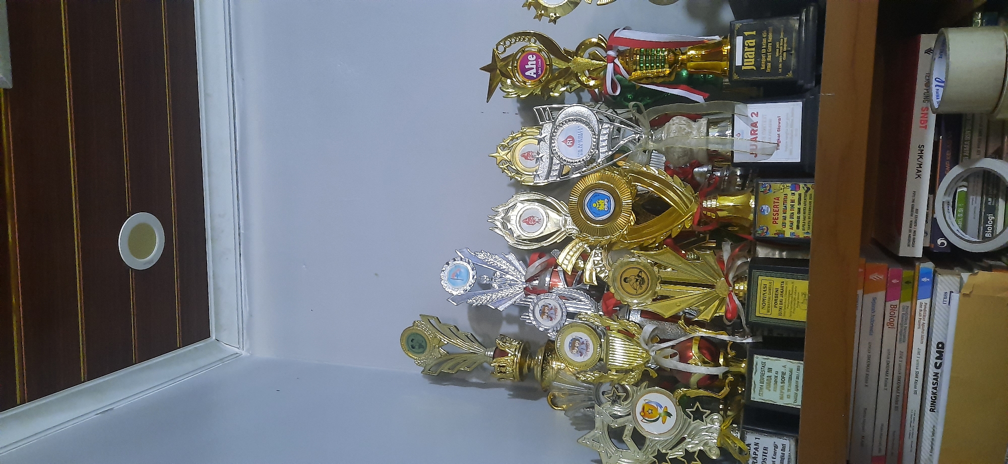 The image displays a collection of trophies and awards displayed on a shelf. The trophies are of various sizes and designs, predominantly gold and silver in color, with some featuring ribbons and emblems. Several trophies have labels with text, some of which appear to be in Indonesian, indicating awards for participation, first, second, and third place, as well as nominations. Below the shelf, a stack of books is visible, with titles such as "RINGKASAN CMD," "Biologi," and "Sejarah Indonesia." A roll of white tape is also present on top of some of the books. The background consists of a light-colored wall and a ceiling with a wooden paneled design and a circular light fixture. The overall scene suggests a domestic or institutional setting, possibly a home or a school, where achievements are recognized and displayed. The location is identified as Cisauk, Indonesia.