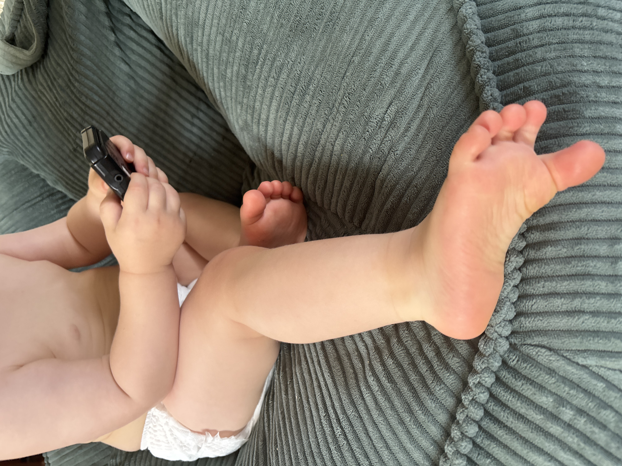 The image shows a close-up of a baby's legs and torso. The baby is wearing a diaper and is sitting on a green, corduroy-textured surface. The baby is holding a black electronic device, possibly a remote control or a small toy, in its hands. The lighting suggests an indoor environment. The image does not provide any information about the location in Naberezhnye Chelny, Russia, or any other specific geographic details.