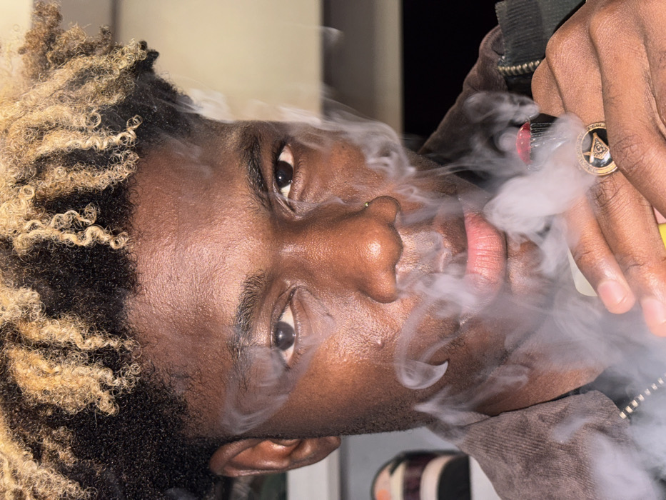 The scene depicts a young individual with dark skin, dark curly hair with bleached blonde or light brown ends, looking directly at the viewer. They are exhaling or producing a dense cloud of white vapor from a vaping device held in their right hand. The device features a red mouthpiece. A ring with a square and compass emblem is visible on the individual's right ring finger. They are wearing a dark, possibly brown, corduroy jacket. A small metal stud is present in their right nostril. The background is dimly lit and out of focus, showing indistinct shapes on a shelf or surface to the left. This event is observed in the city of Igboefon, Nigeria.