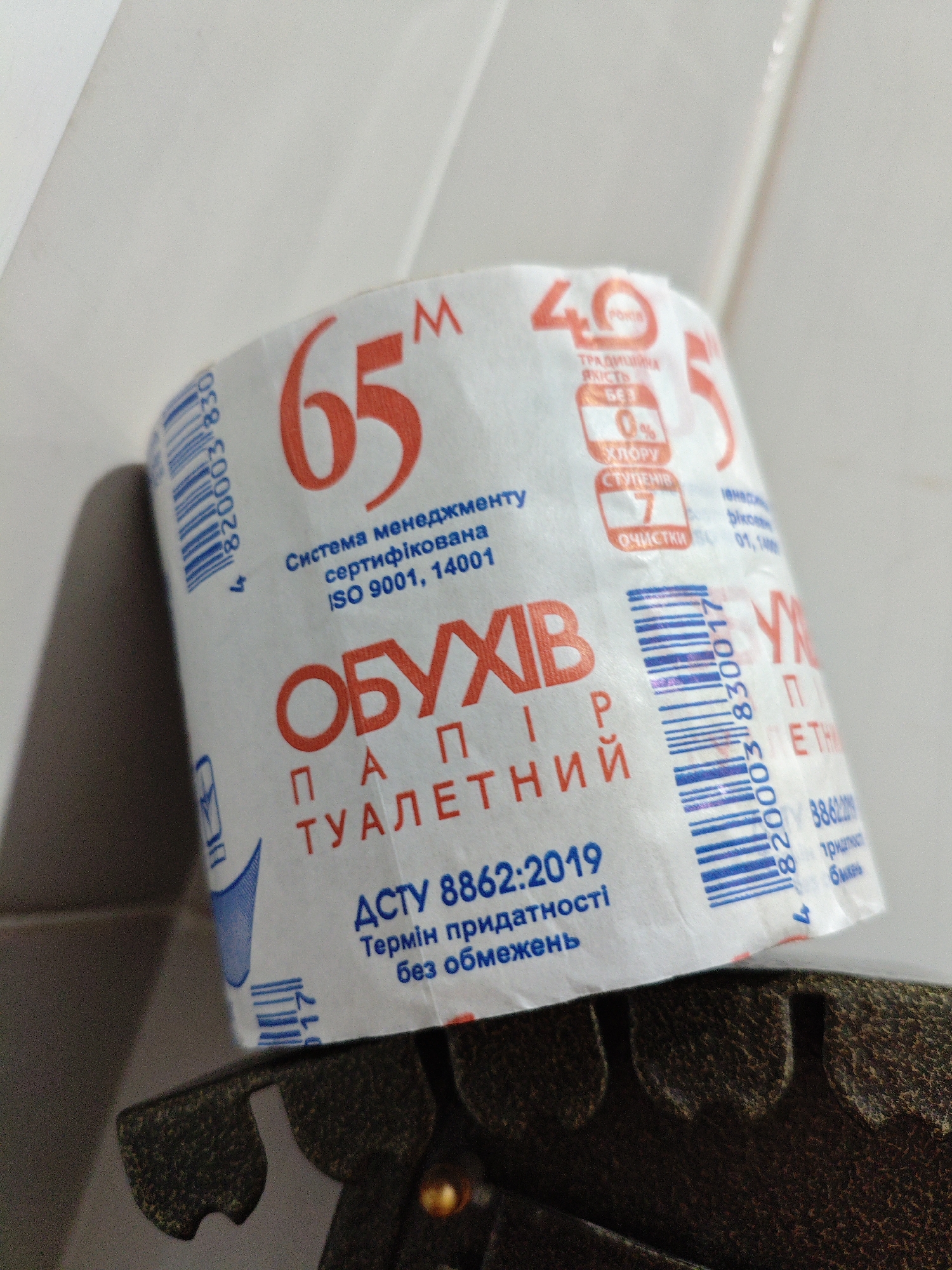 The image shows a roll of toilet paper with Ukrainian text on it, placed on a dark, textured surface. The toilet paper roll is partially unrolled, revealing the branding and product information. The text on the packaging is primarily in red and blue. Prominent features include the numbers "65" and "40" in large red font, and the words "ОБУХІВ ПАПІР ТУАЛЕТНИЙ" (Obukhiv Paper Toilet). There are also barcodes and smaller text details, some of which are difficult to read due to the angle and lighting. The background is a plain, light-colored surface, possibly a wall. The image is taken from a close-up perspective, focusing on the product. There are no people or animals present in the image. The environment appears to be indoors, likely a household or retail setting. There is no indication of the specific location in Kyiv, Ukraine.